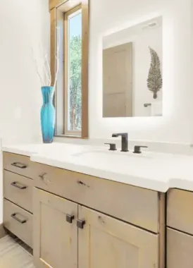Bathroom vanity with light-up mirror, blue vase, wooden cabinets, and a window with a view by Syn-Mar