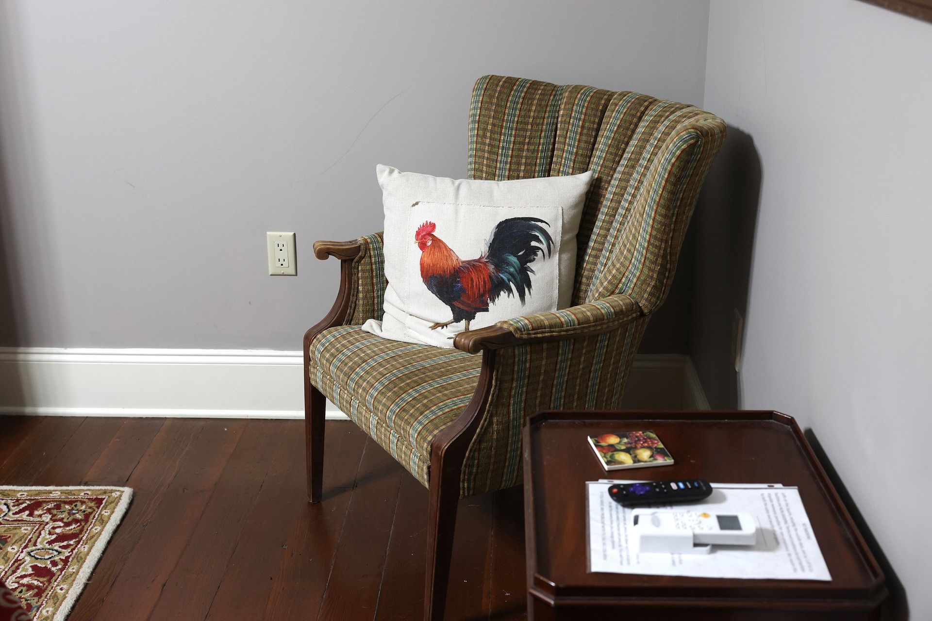 A chair with a rooster pillow on it is in a living room next to a table.