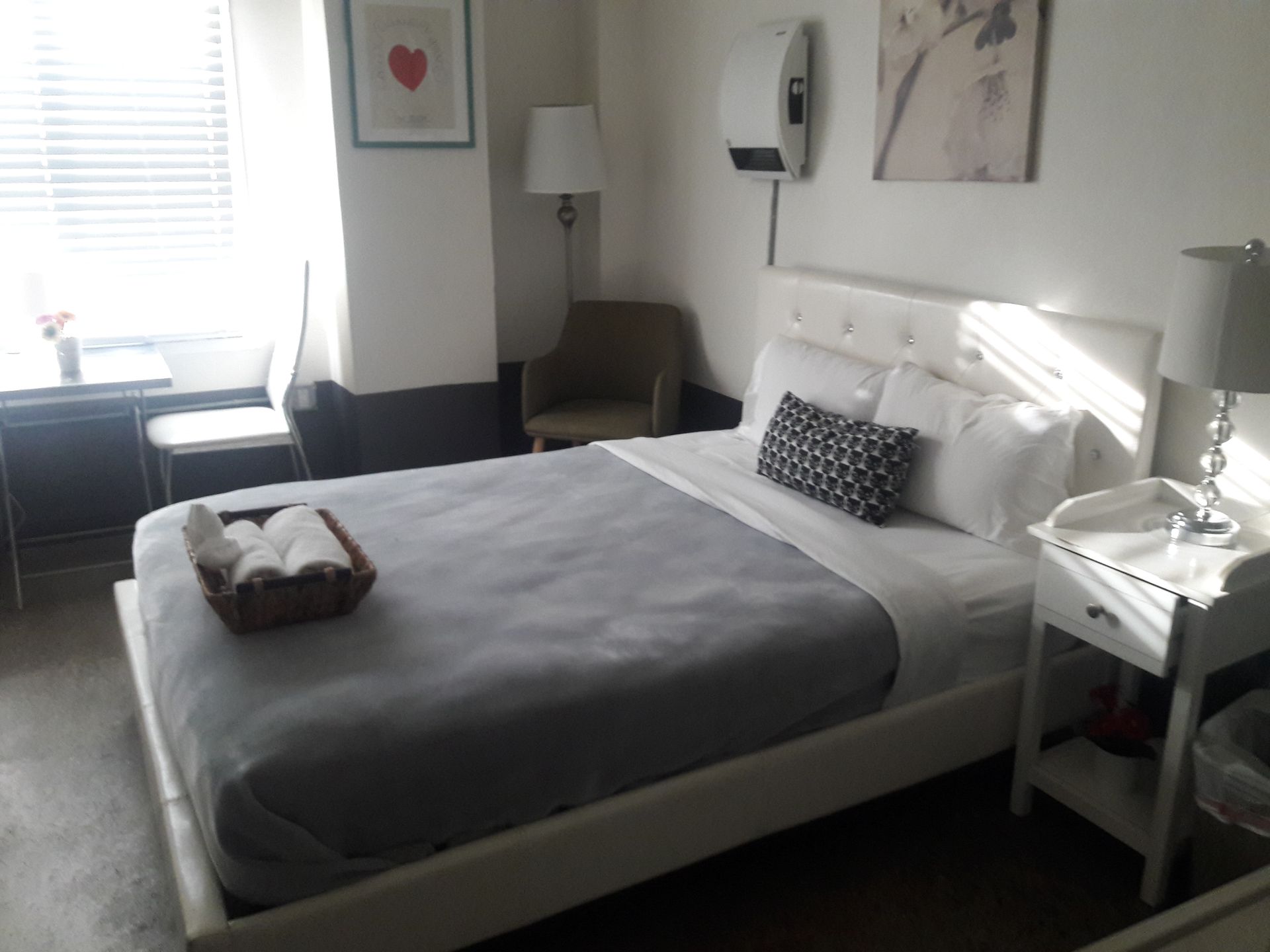A hotel room featuring a bed with a gray blanket and a basket of towels, a nightstand, an armchair, and a wall heater.