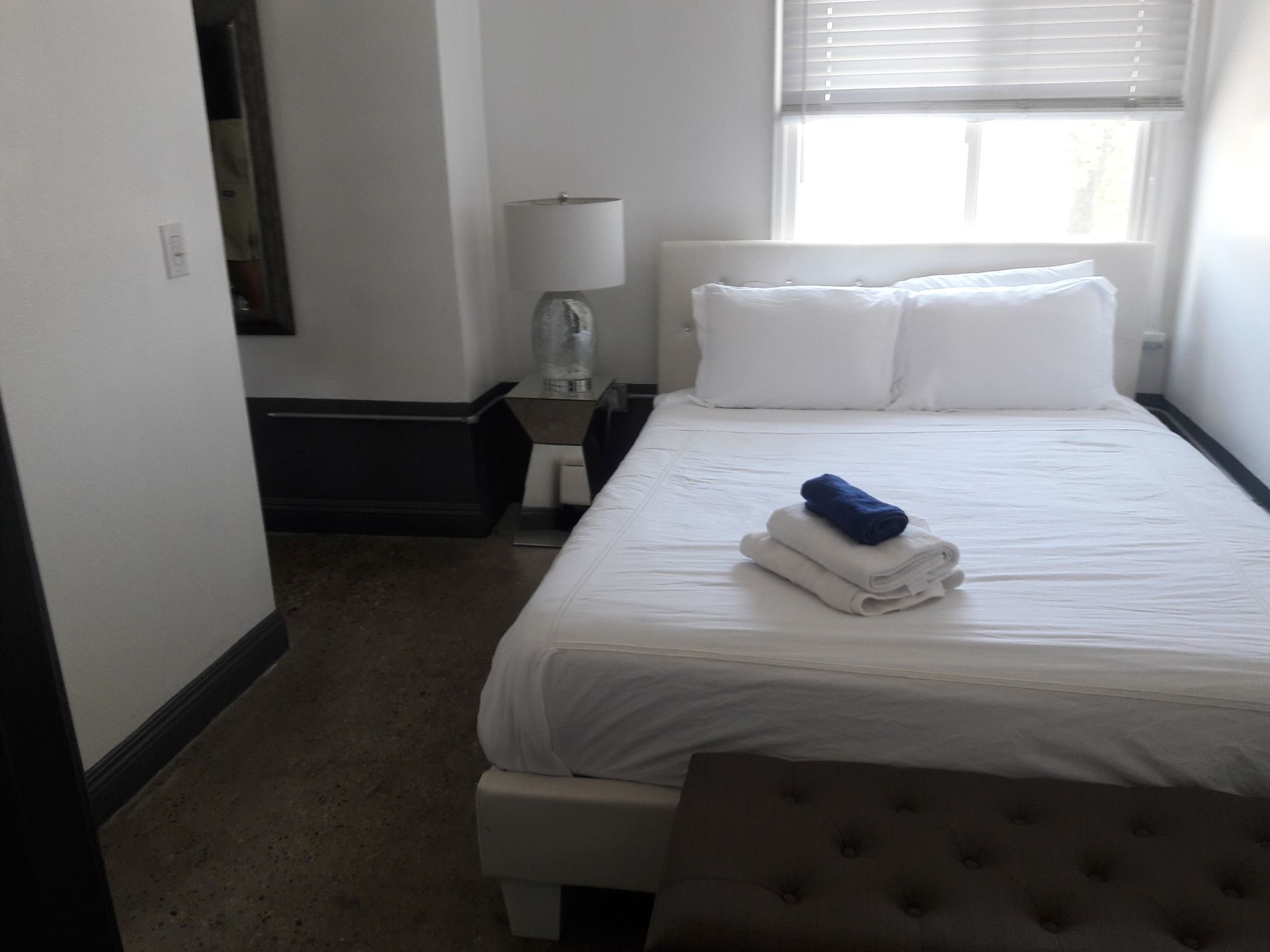 A hotel bedroom featuring a white-made bed with folded towels, a bedside lamp, and a window with blinds.