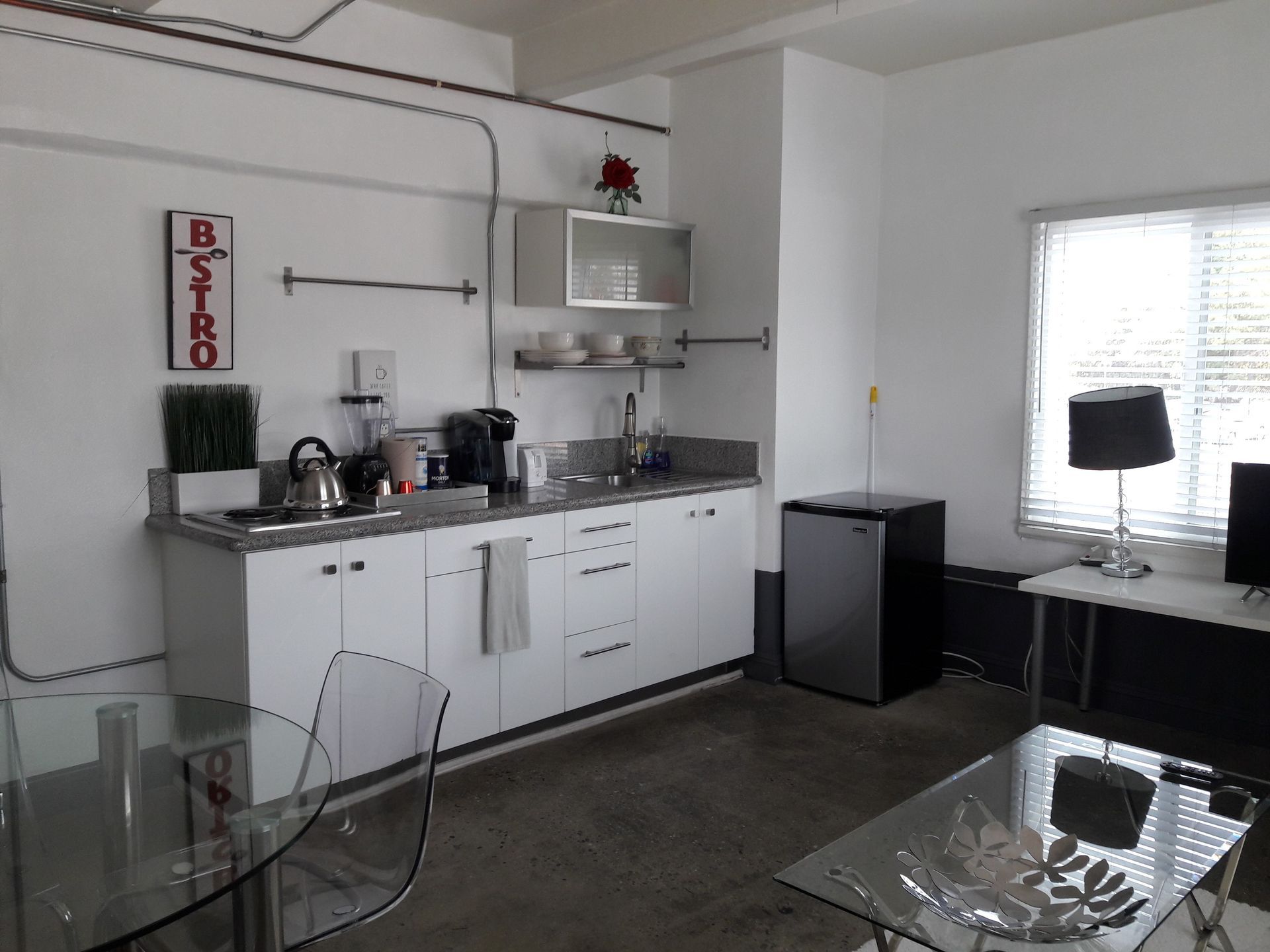 A kitchen area with white cabinets, a small refrigerator, a glass dining table, and a window with blinds in a bright room.