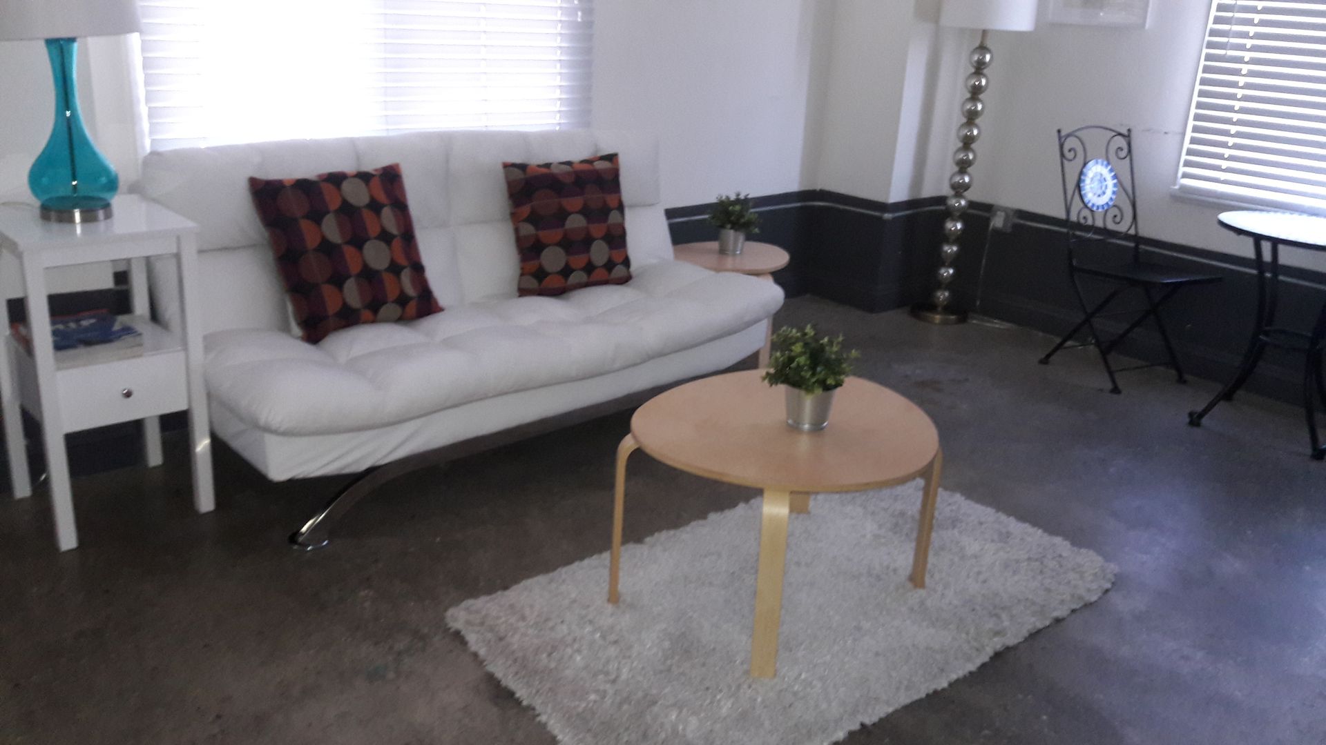 A white sofa with two patterned pillows, a round wooden coffee table on a white rug, and a side table on concrete flooring.