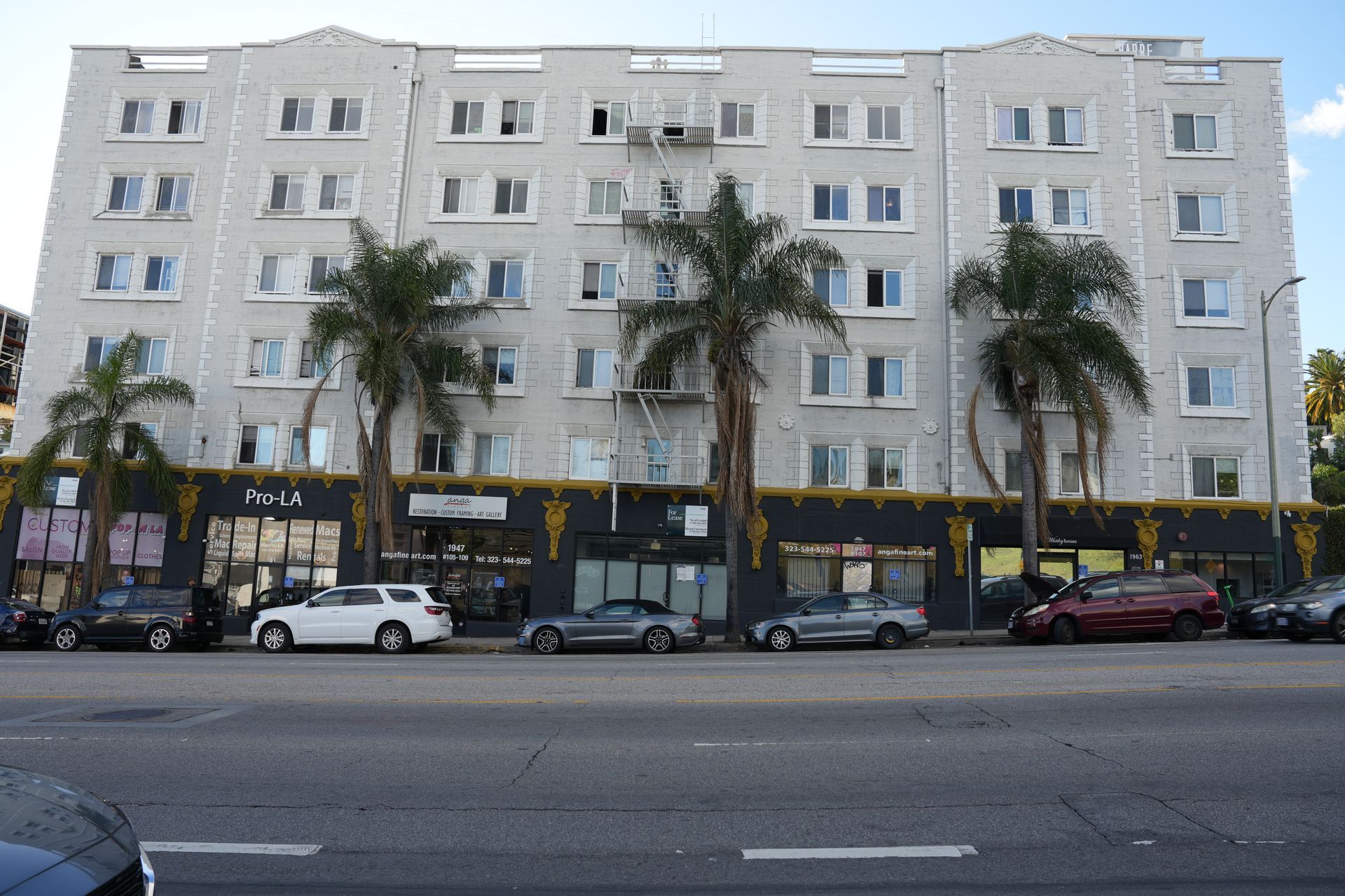 A five-story white apartment building with storefronts on the ground level, featuring three tall palm trees in front.