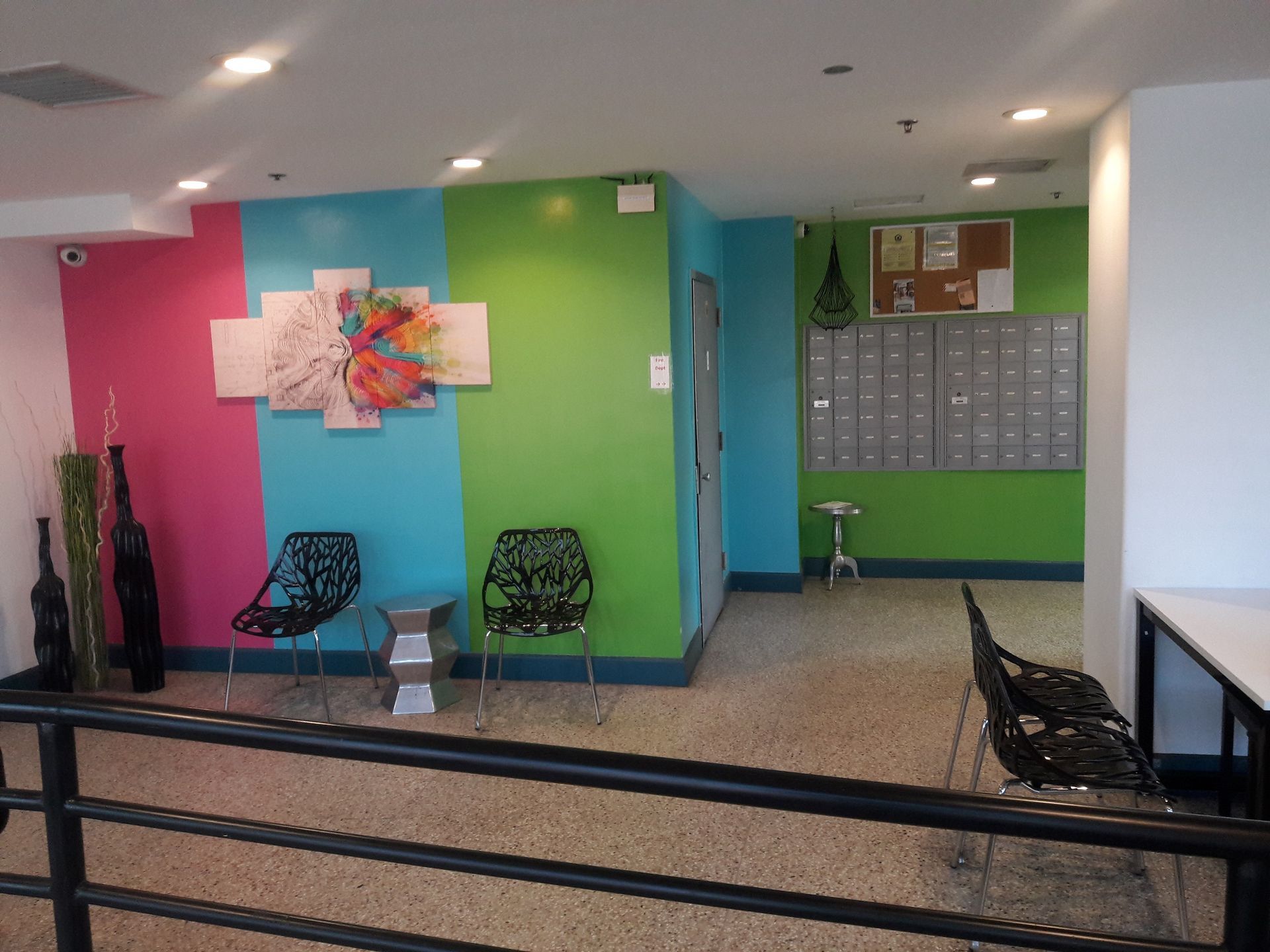 A lobby with a colorful wall of pink, blue, and green, black wire chairs, and a mail area with a corkboard.