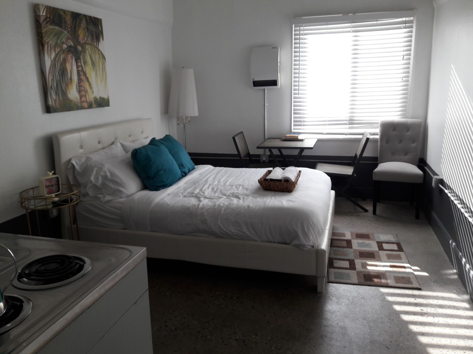 A studio hotel room with a queen bed, two blue pillows, a small dining table, a chair, and a view of a window.
