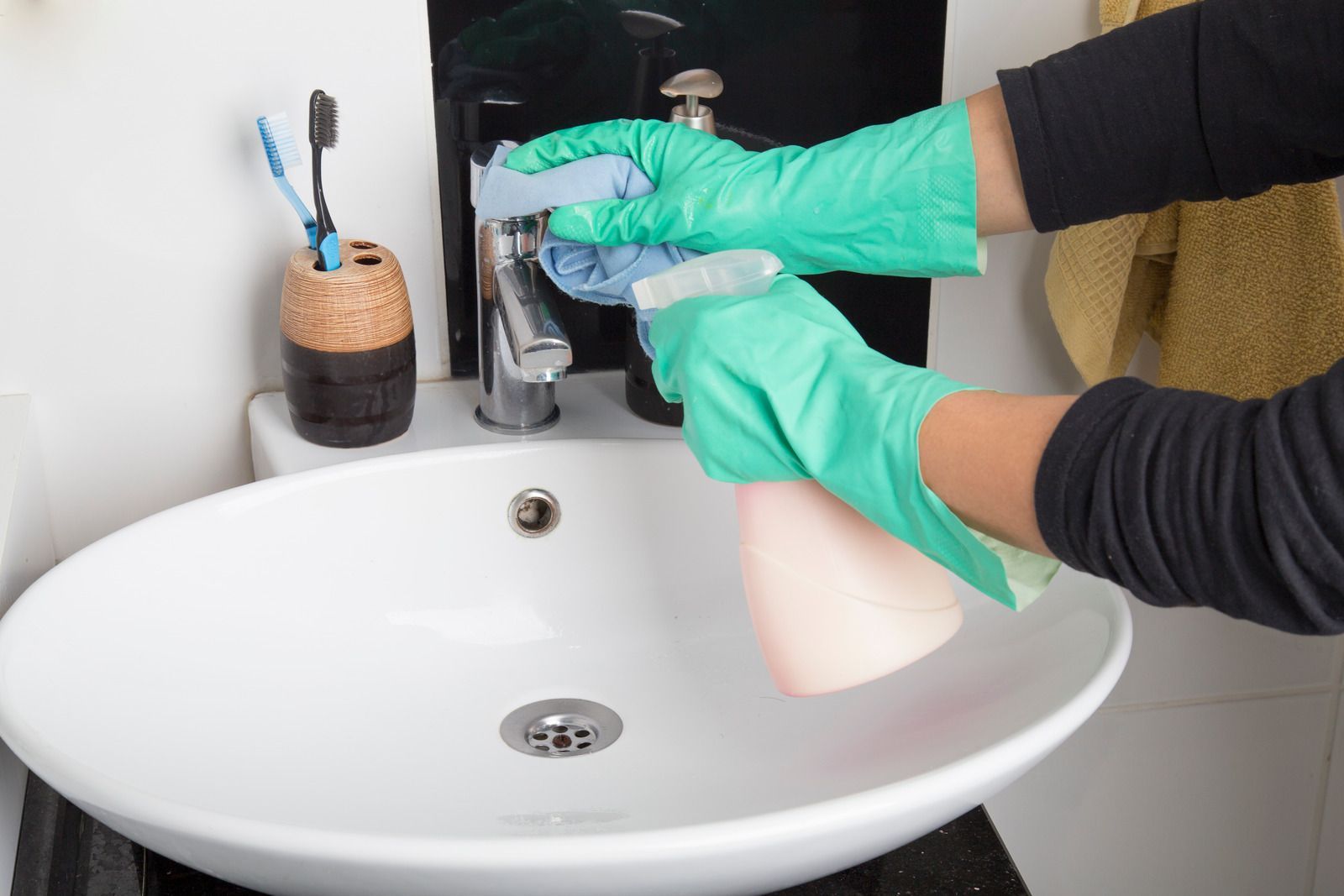 Person wearing green gloves sprays and wipes a bathroom faucet and sink.