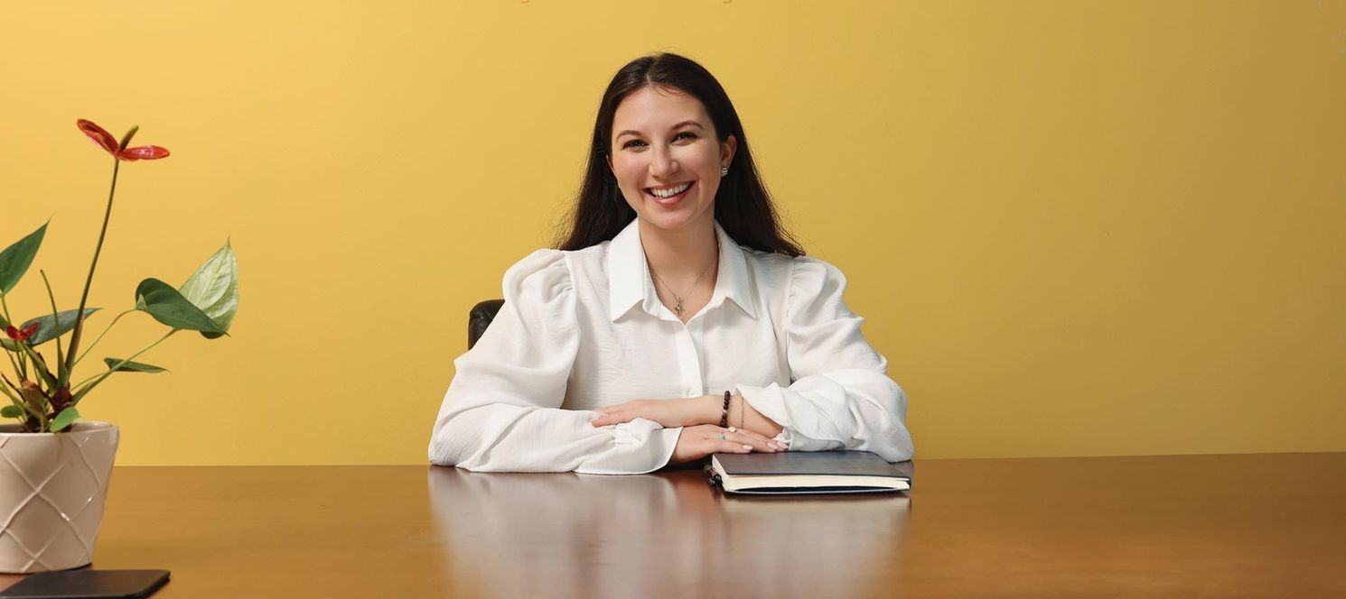 Woman in white shirt smiles, sitting at a brown desk with a potted plant against a yellow background.