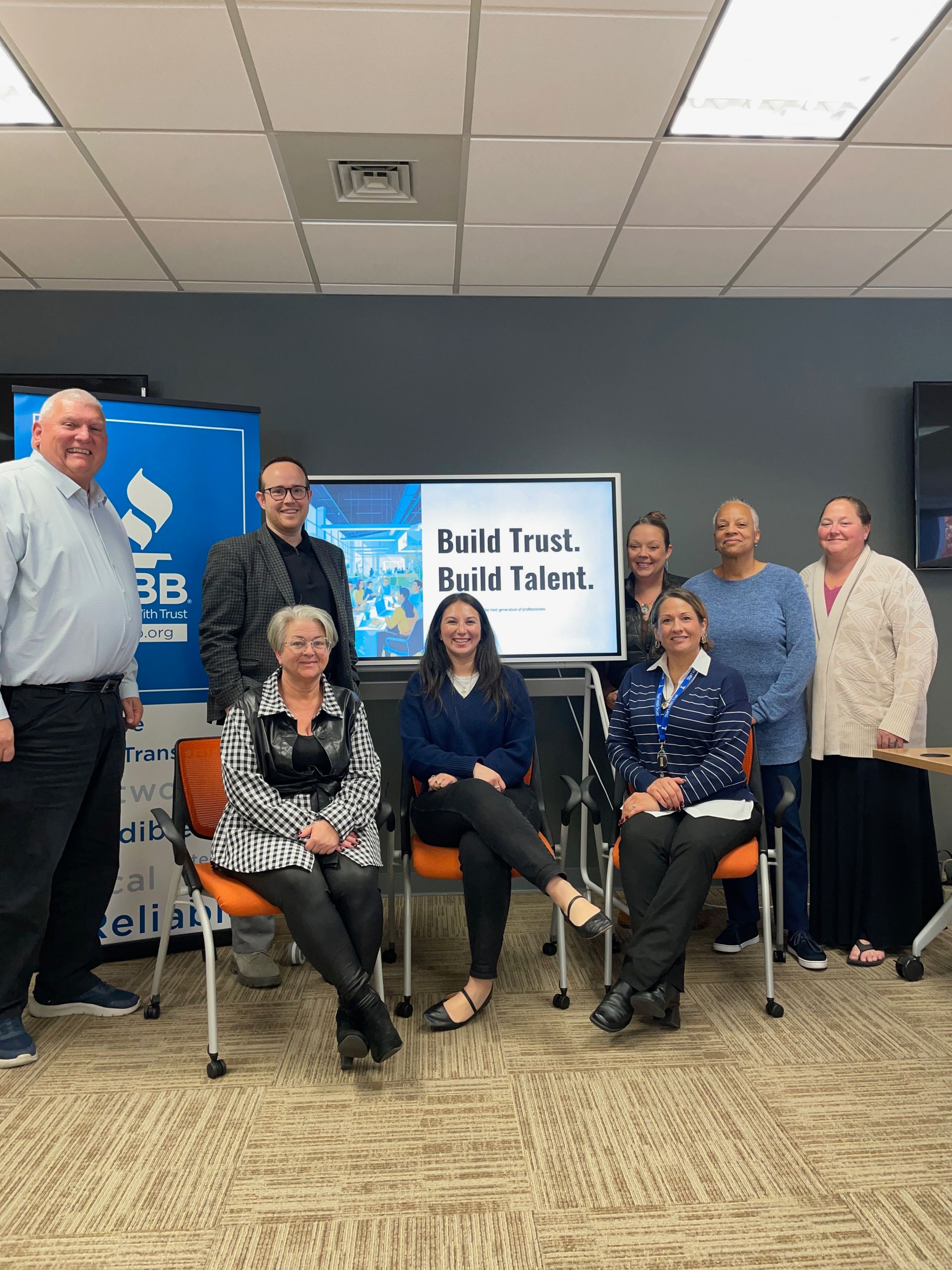 Woman giving a presentation at a table, others seated and listening. Meeting room setting.