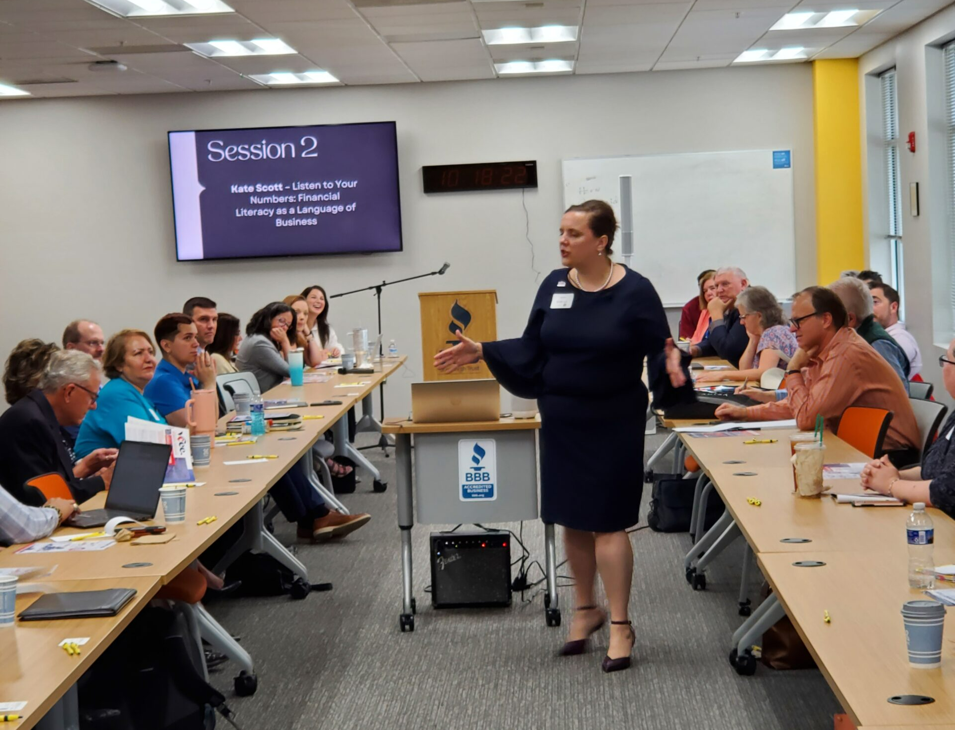 Woman giving a presentation in a conference room. Audience seated at tables, listening.