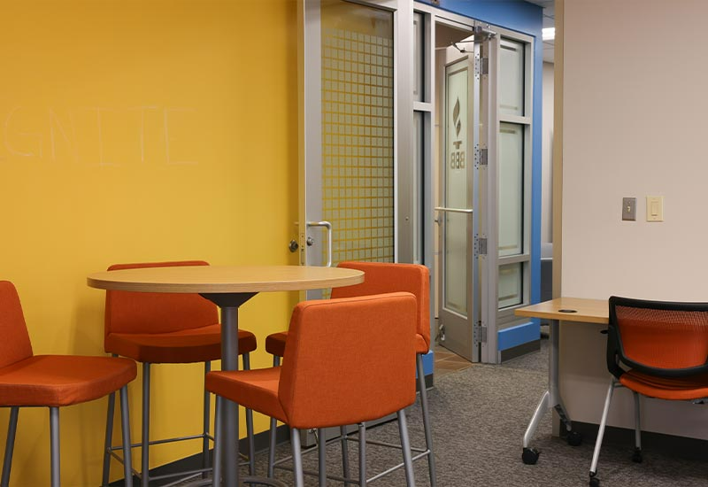 A brightly-lit room with orange chairs and a table, leading to a blue-framed doorway.