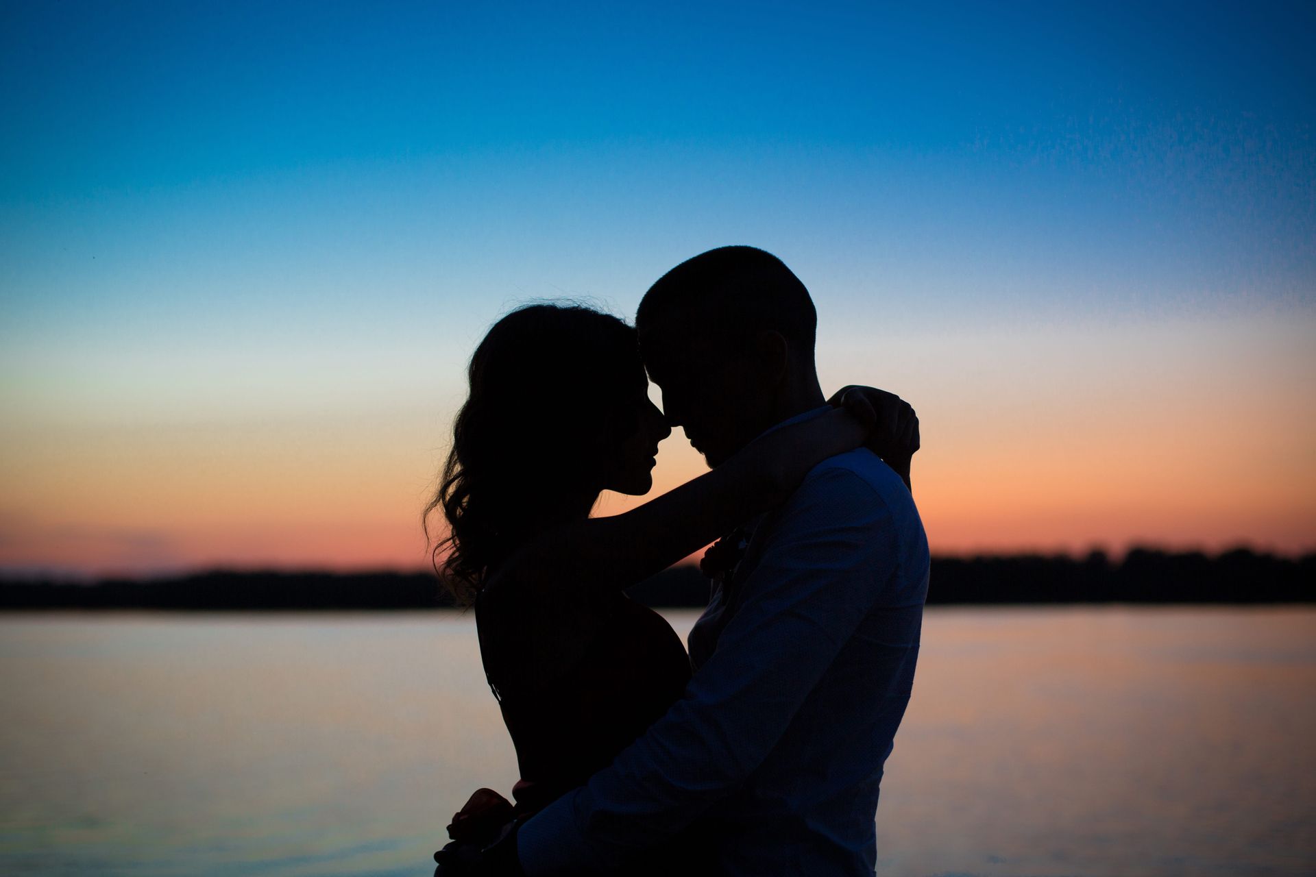 Silhouetted couple embracing by a calm lake at sunset, with a blue-orange sky