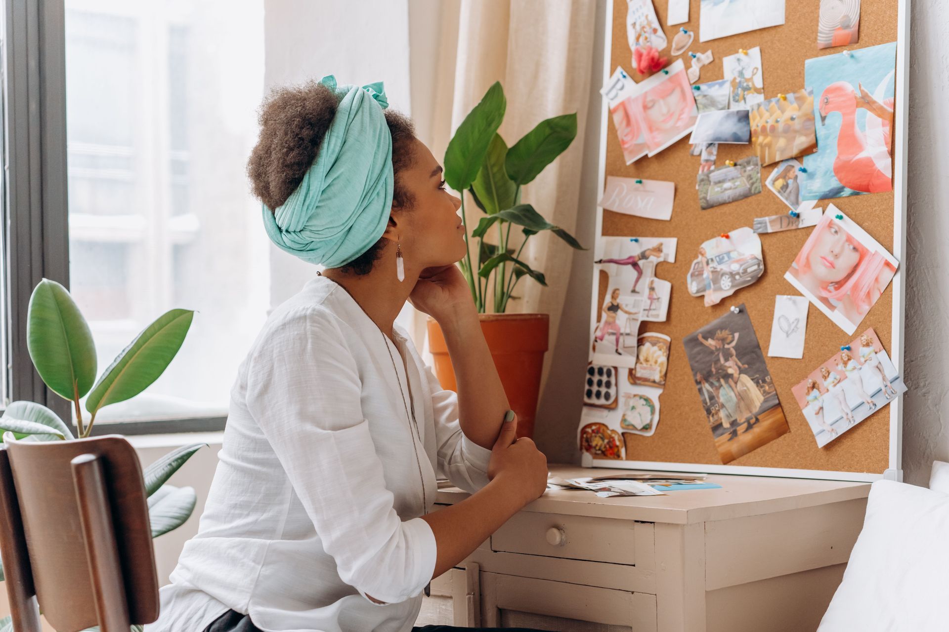 Woman pinning notes on a corkboard in a bright home office, holding a plant.