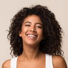 A woman with curly hair is smiling and wearing a white tank top.