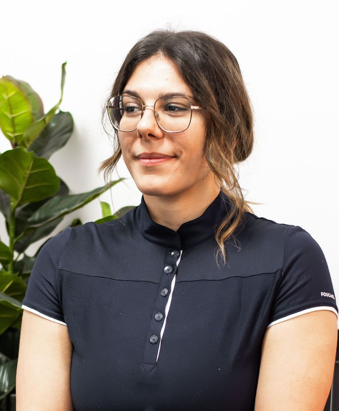 A woman wearing glasses and a blue shirt is sitting in front of a plant.