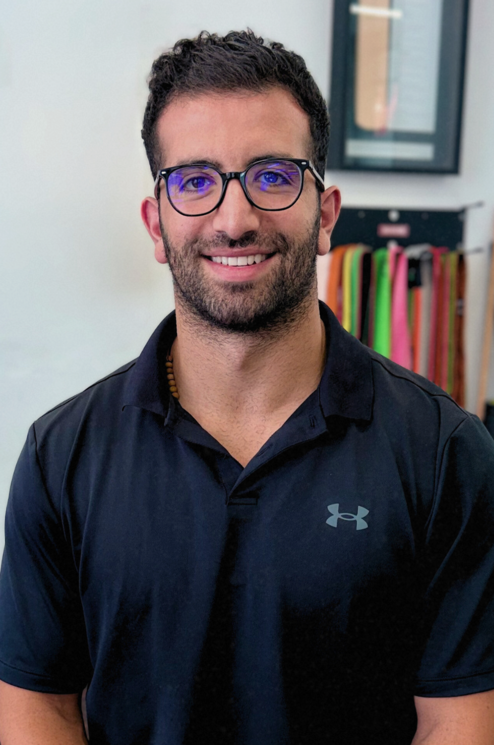 Man wearing glasses and black shirt smiles, standing in front of a white wall.