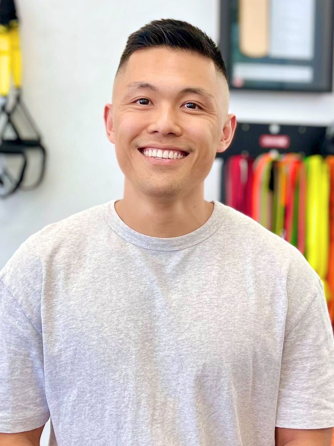 Man with short hair smiles at the camera, wearing a light gray t-shirt. Interior setting, with equipment visible.