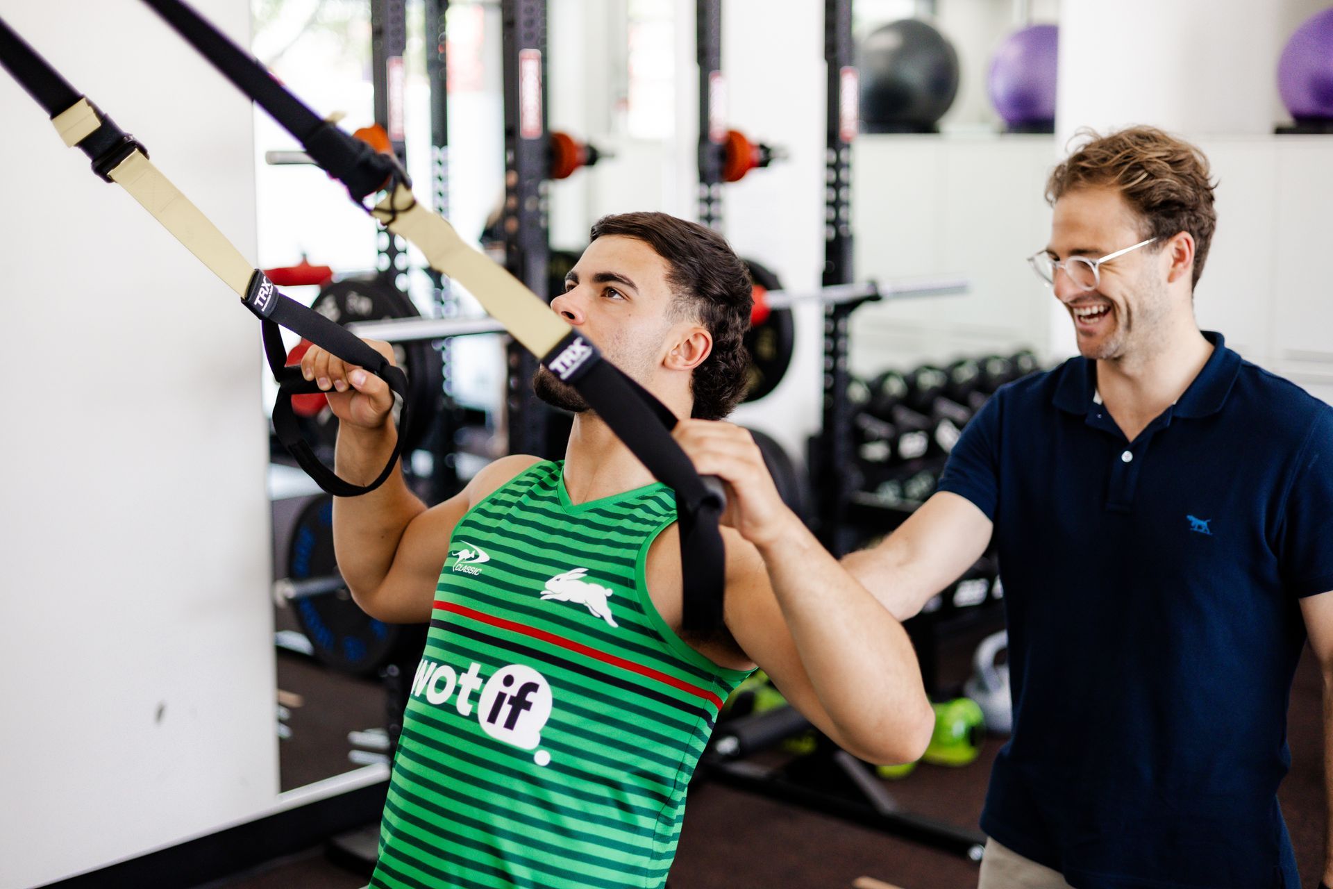 Man using TRX straps, assisted by another man in a gym setting. The man is wearing a green shirt and focused.