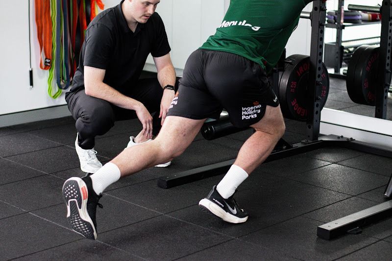 A man is helping another man stretch his legs in a gym.