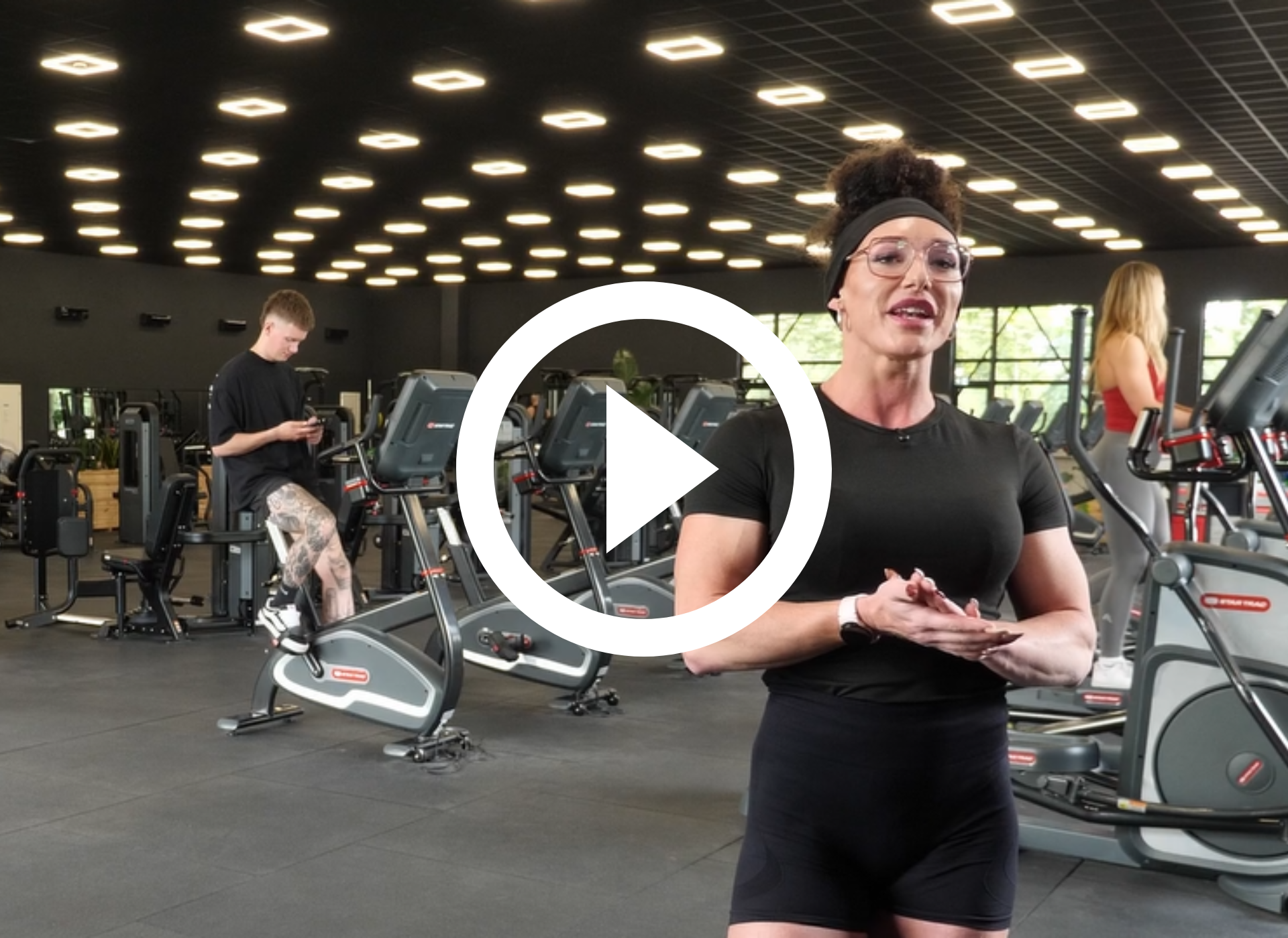 Vrouw in sportschool, zwart tanktopje, pratend. Fitnessapparatuur op de achtergrond.