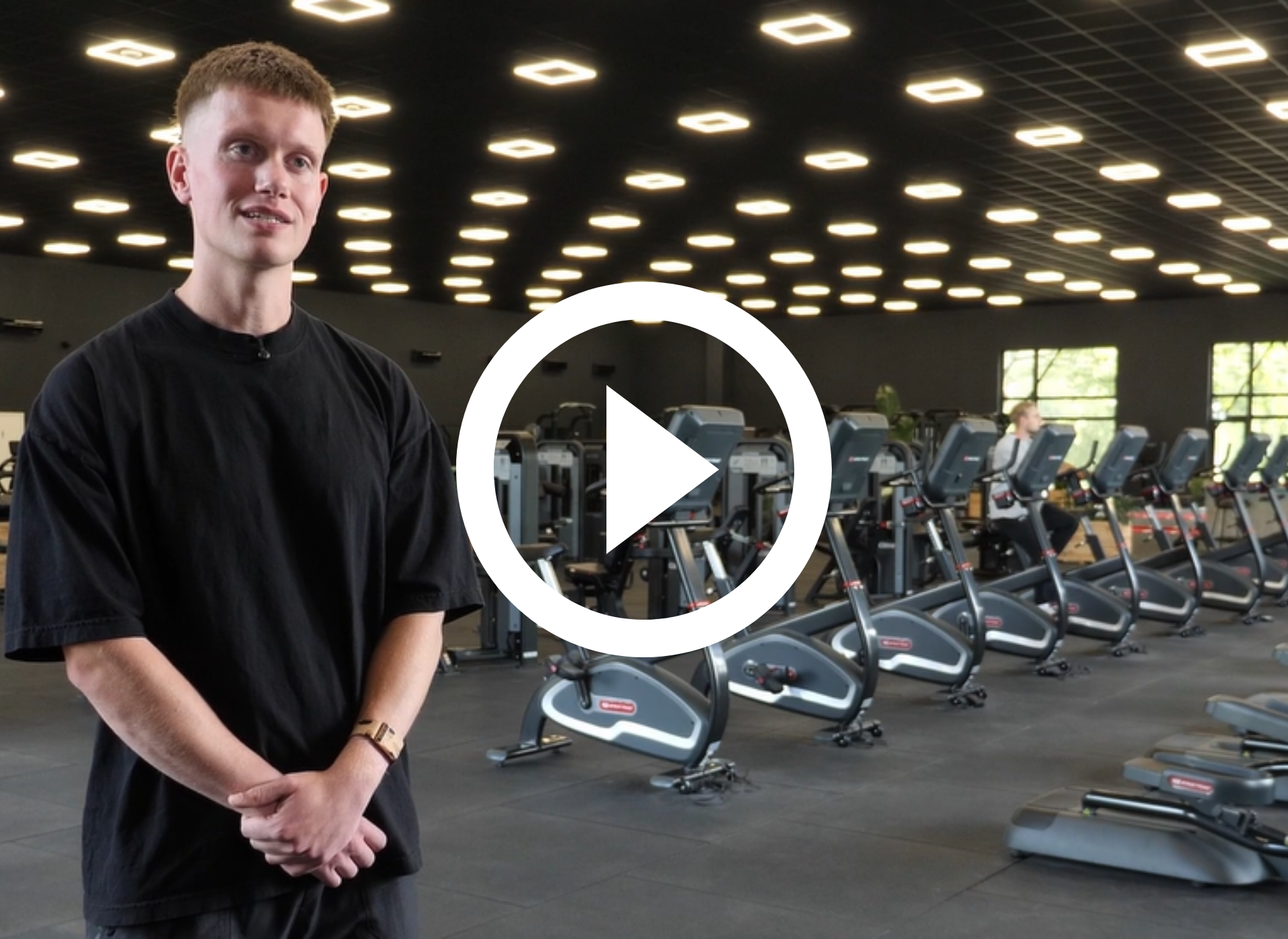 Man in grijs shirt in een sportschool, met hometrainers achter zich. Hij kijkt in de camera.