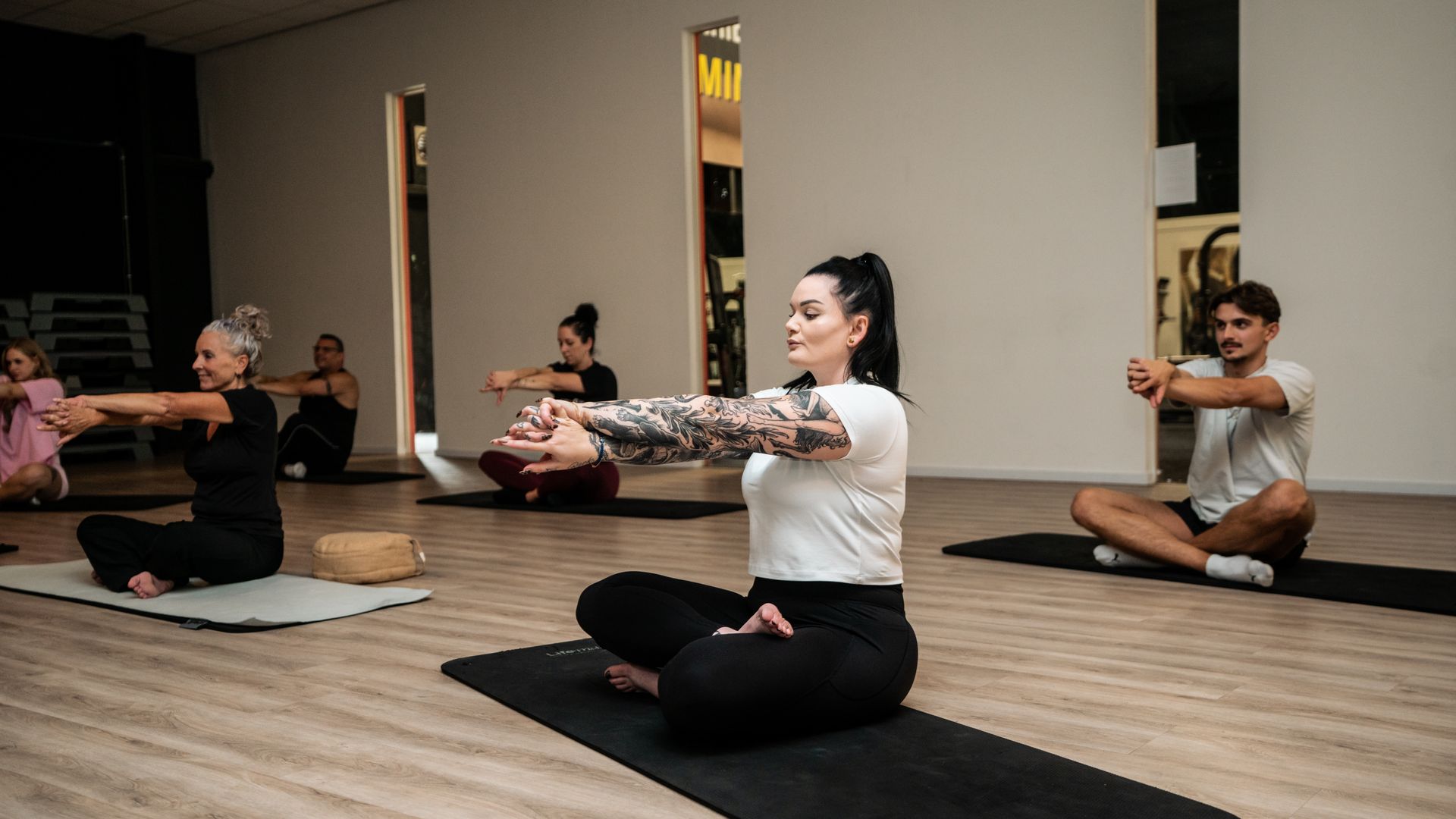 Een vrouw in sportkleding strekt zich uit op een roze mat in een studio met andere vrouwen.