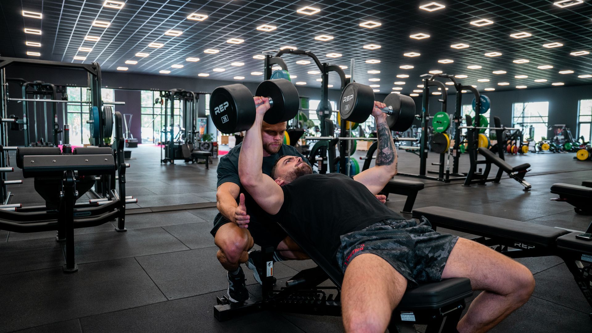 Personal trainer begeleidt een man bij het doen van push-ups in een sportschool. De trainer draagt een zwart-oranje broek; de man draagt een zwarte broek.