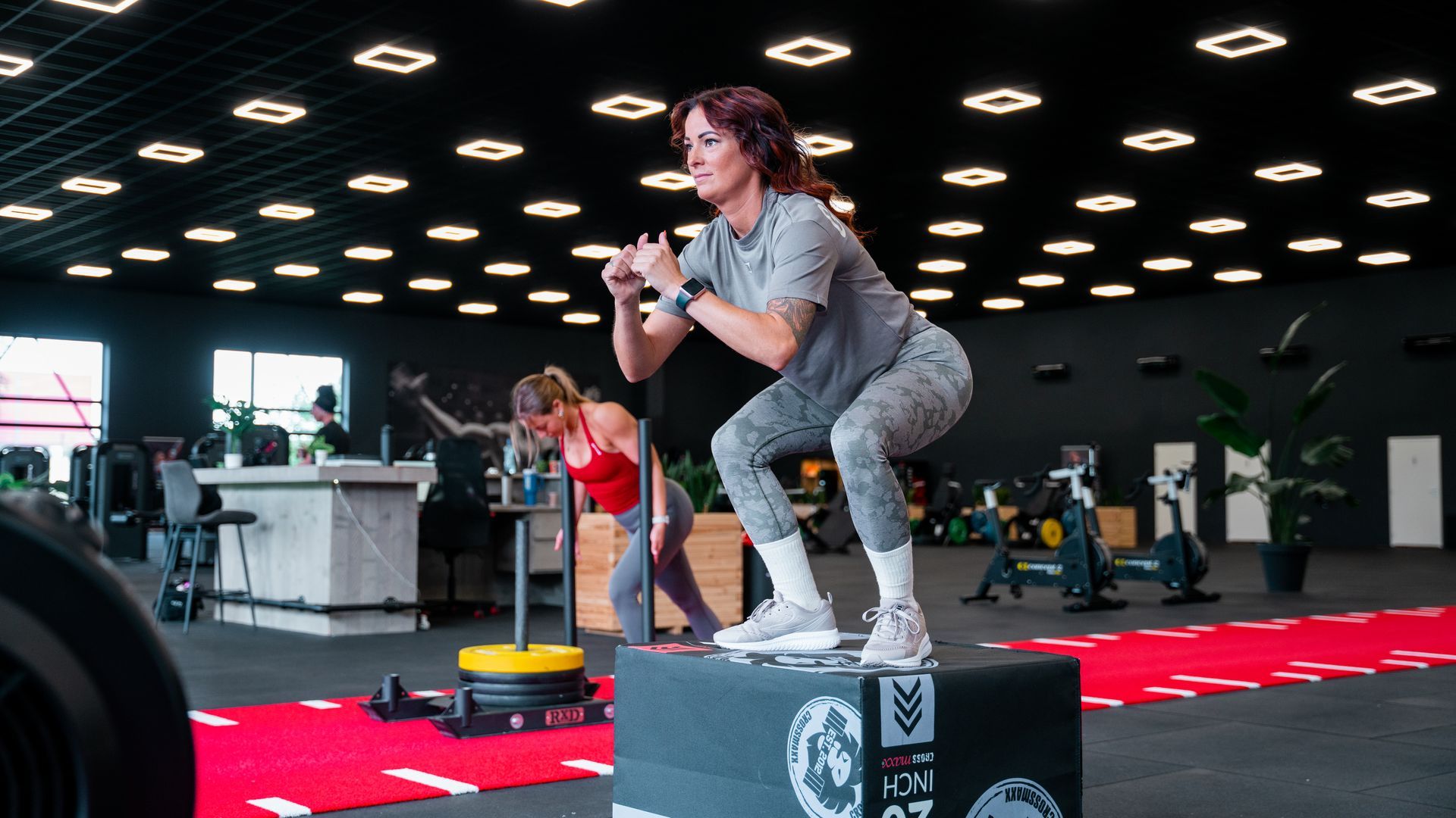 Twee vrouwen in zwarte danspakken zitten op een yogamat. De een geeft de ander instructies en gebaart in een dansstudio.