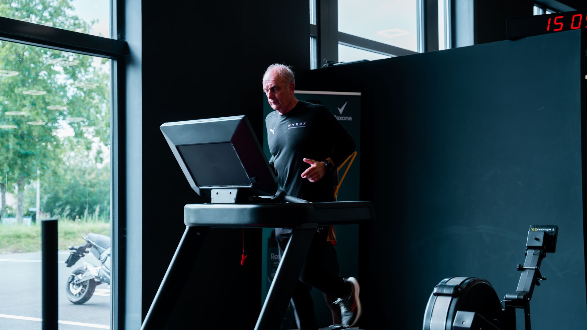 Een man rent op een loopband in een sportschool, naast twee anderen, met een groot raam op de achtergrond.