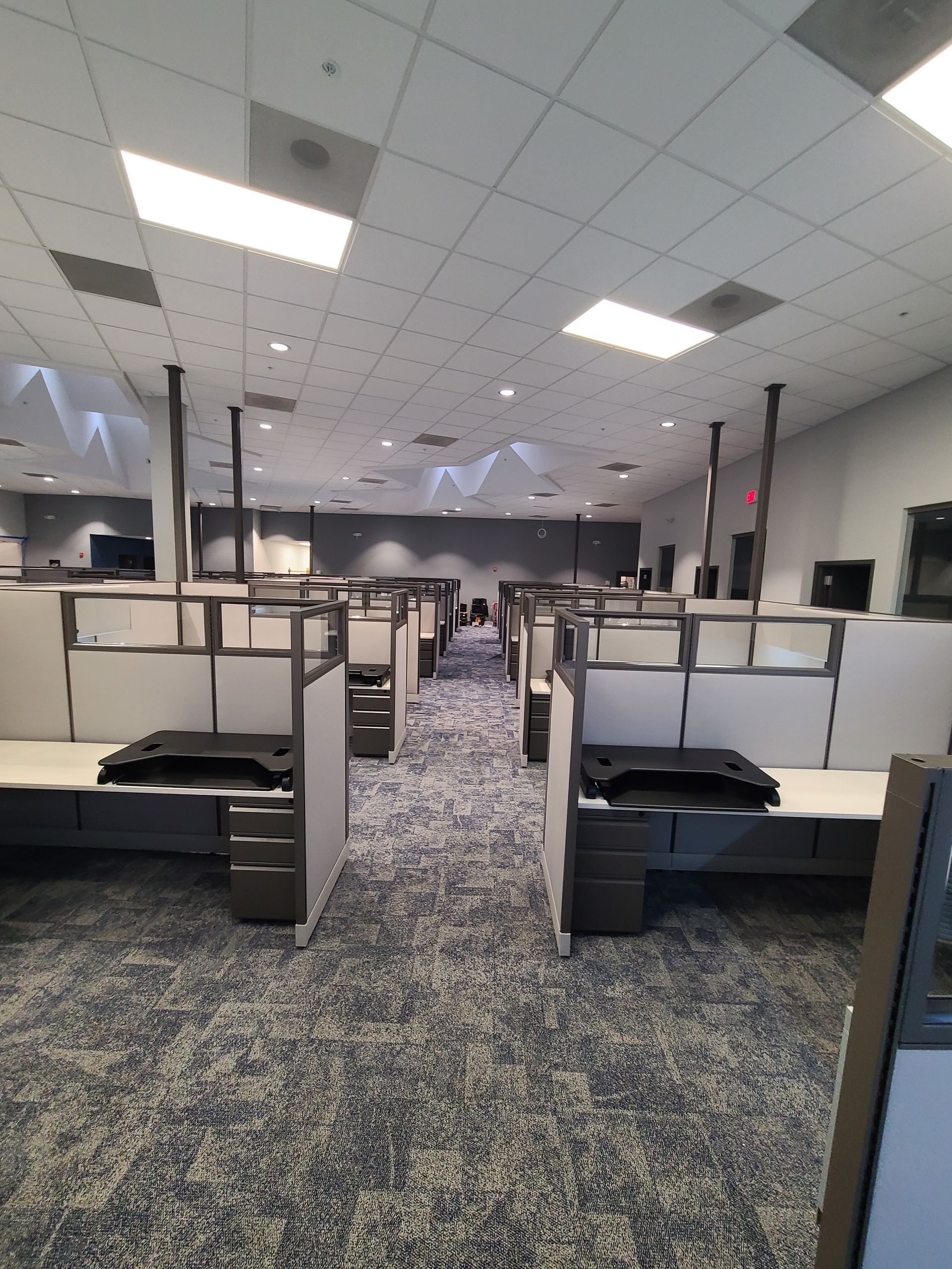 Office cubicles in rows, gray carpet, white ceiling with rectangular lights.