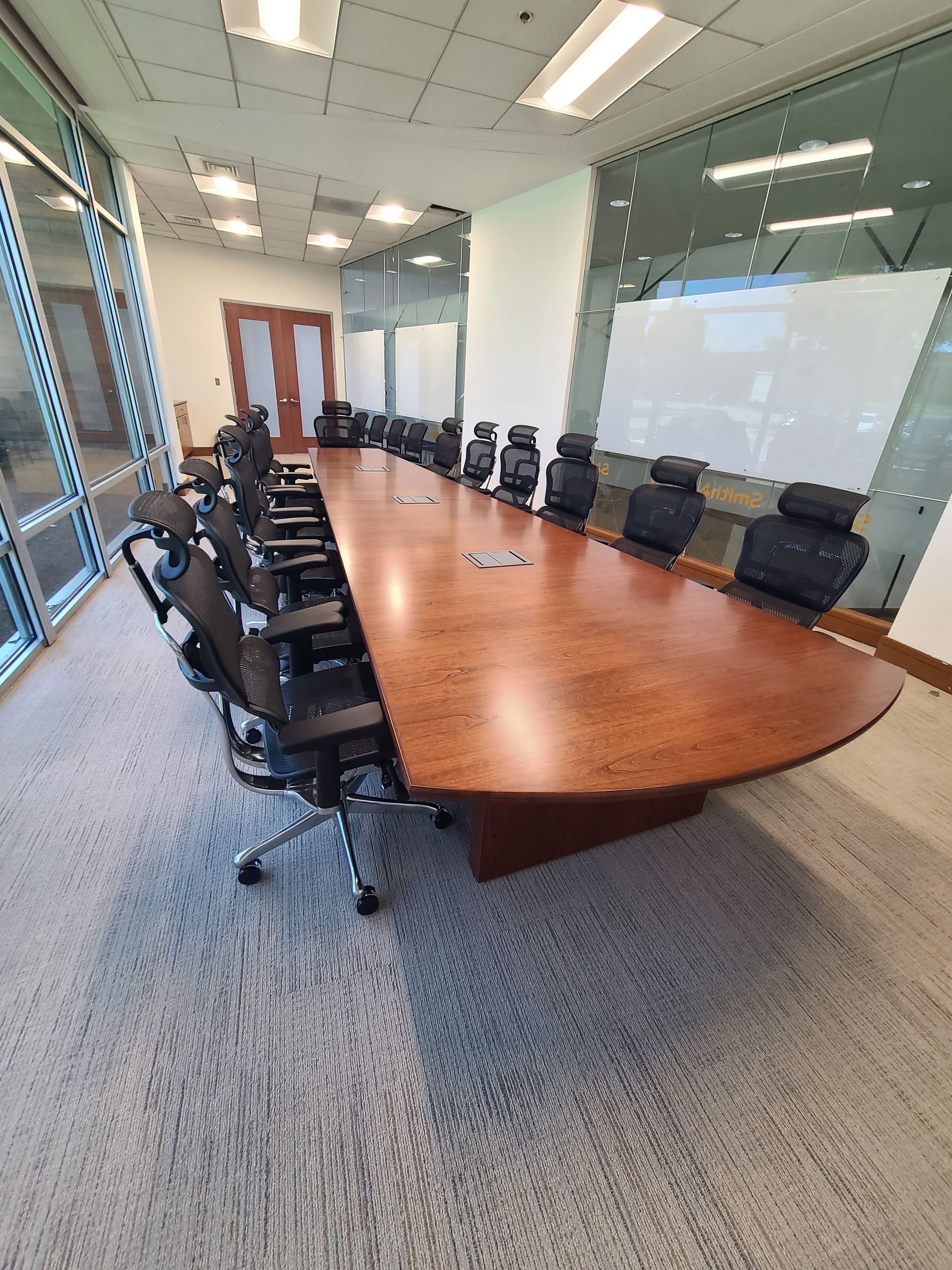 Long wooden conference table with black chairs in a room with windows and whiteboards.