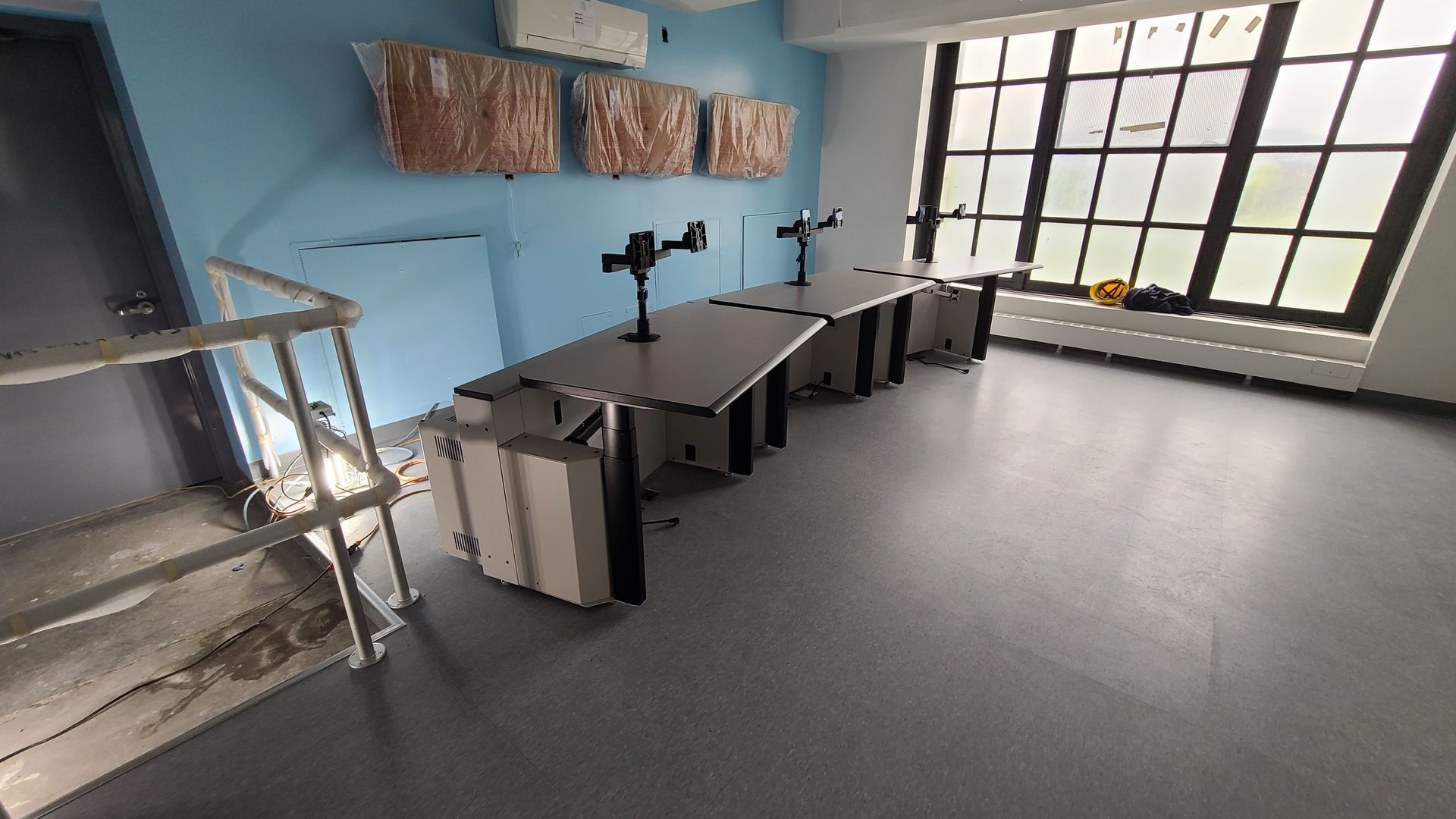 An empty office with a long desk, monitors, and large window. Blue walls and grey floors.
