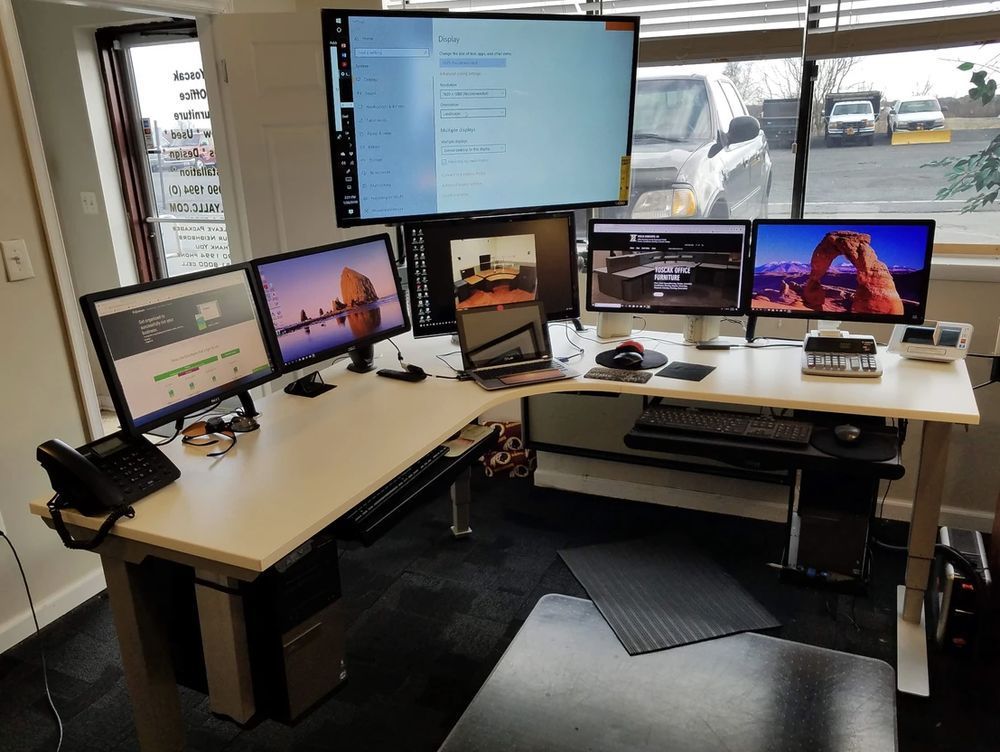 L-shaped desk with multiple monitors displaying websites and software; a laptop is open; the setting appears to be an office.