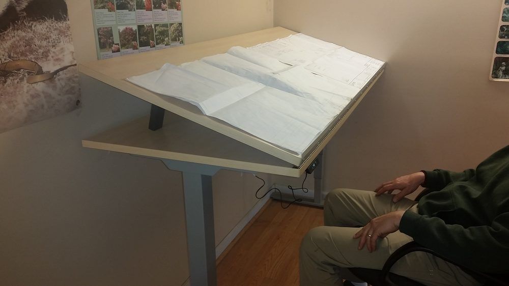 Person seated at a tilted drafting table with a drawing on it.  Table is in a corner, with a drawing on it.
