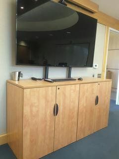 A large TV mounted above a light wood cabinet with three doors, in an office setting.