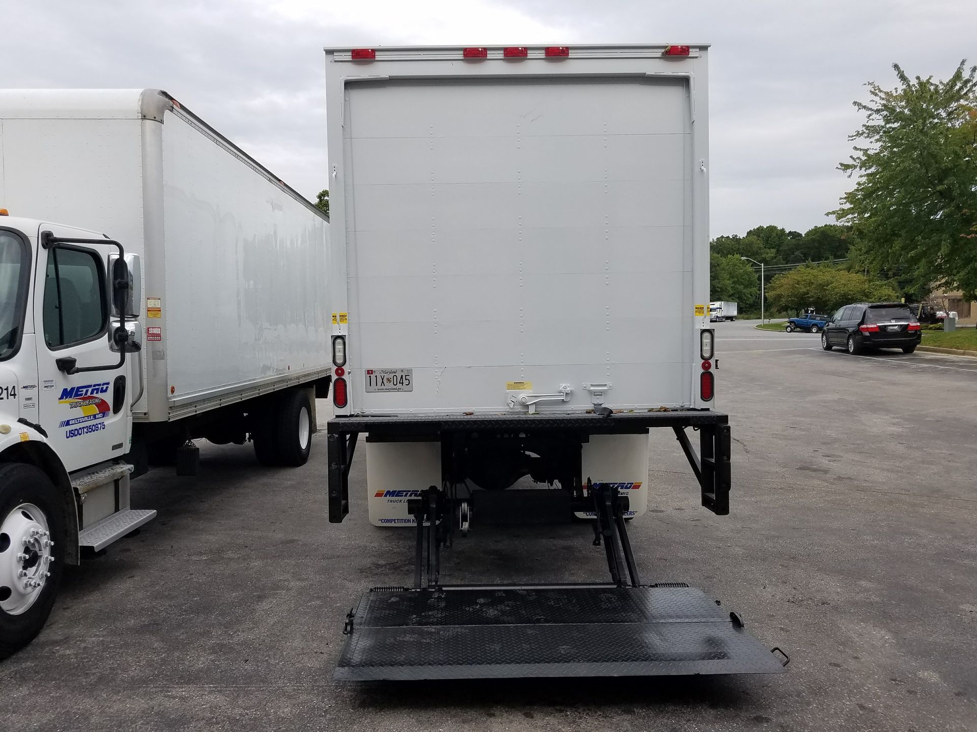 Rear view of a white box truck with a lift gate, parked on pavement. Another truck is partially visible to the left.