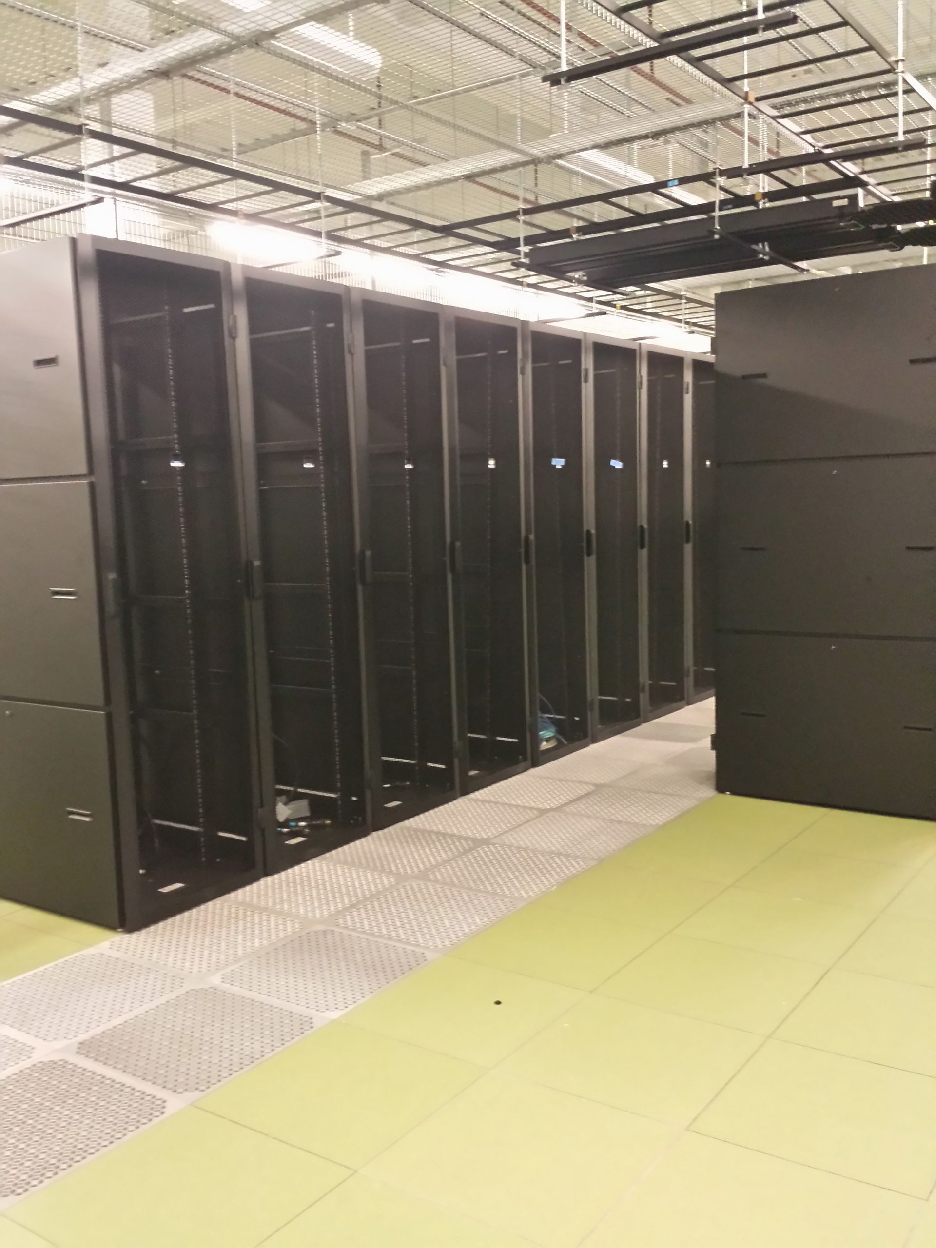 Row of black server racks in a data center; green and gray flooring, industrial ceiling.