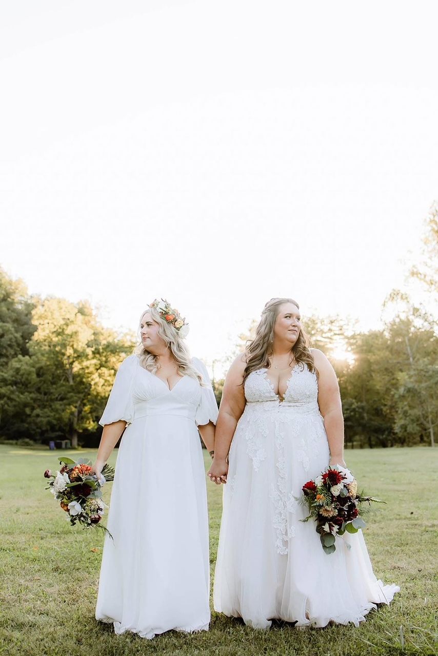 Two brides are standing next to each other in a field holding hands.