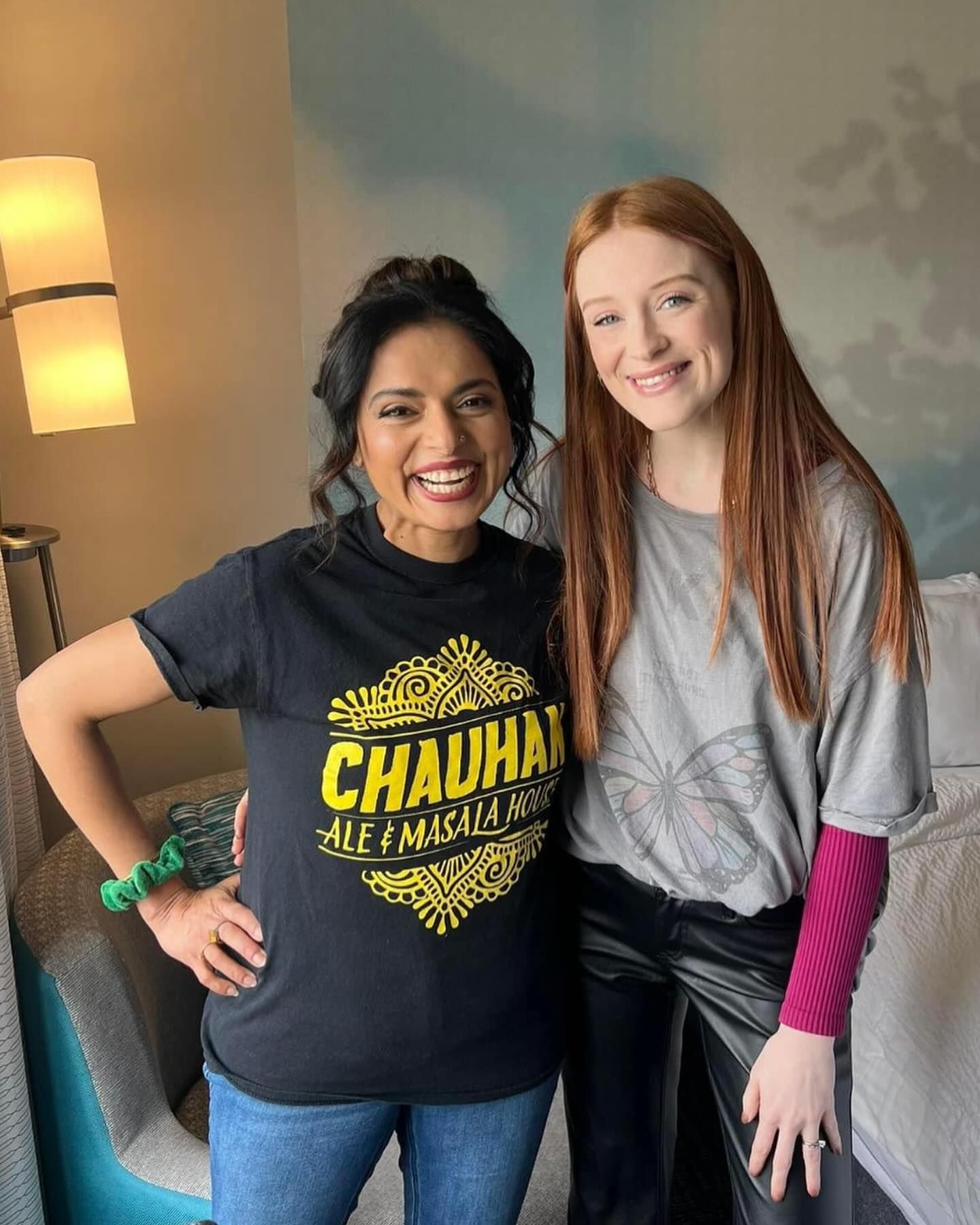 Two women are posing for a picture in a hotel room . one of the women is wearing a chauhan shirt.