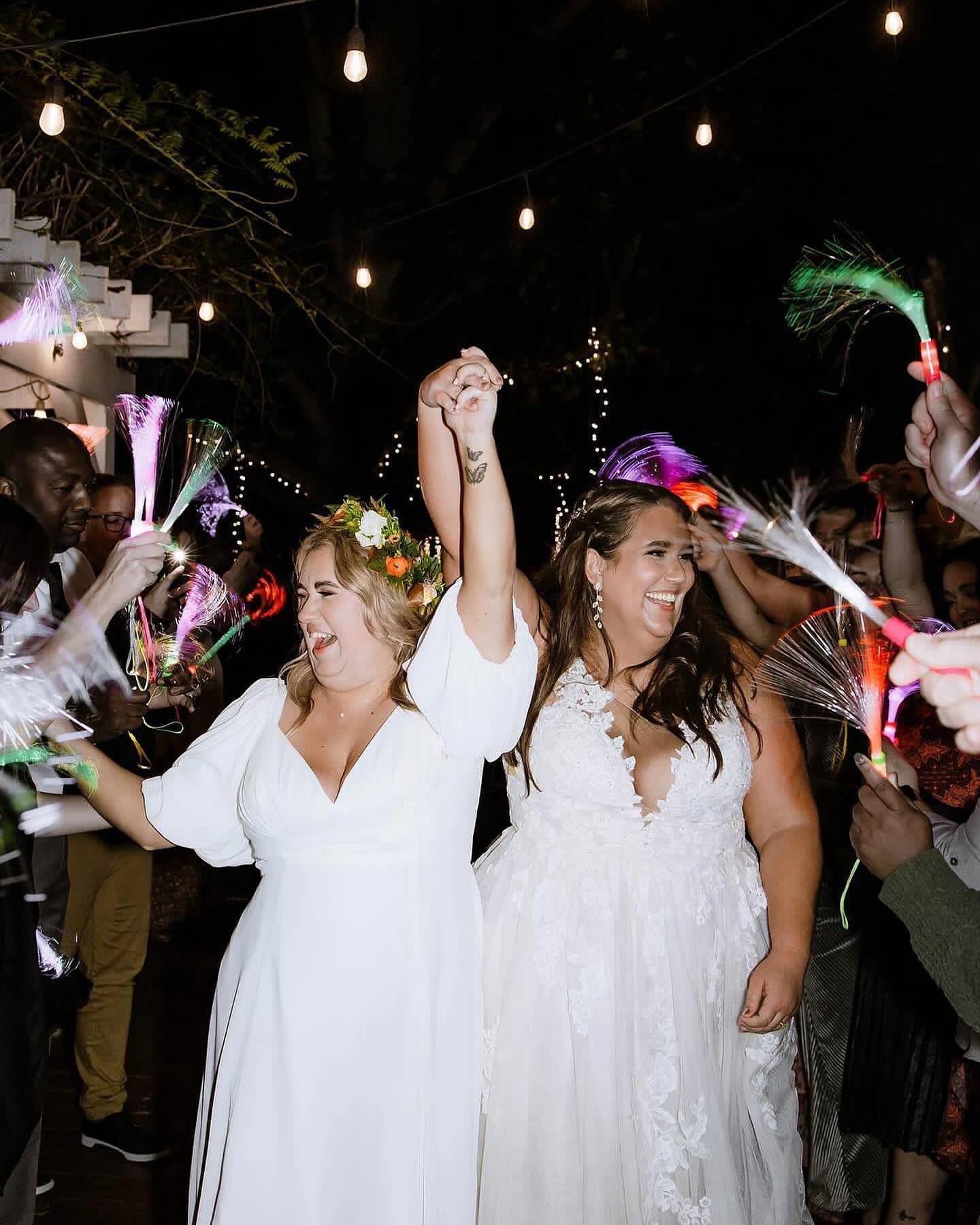 Two women in white dresses are holding hands and dancing at a wedding reception.