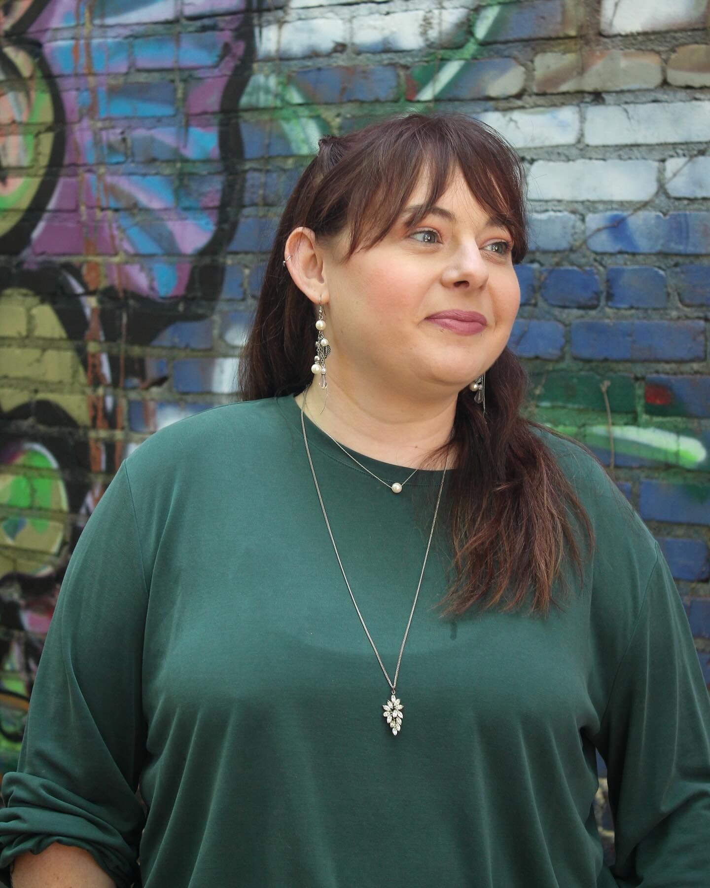 A woman in a green shirt is standing in front of a brick wall with graffiti on it.