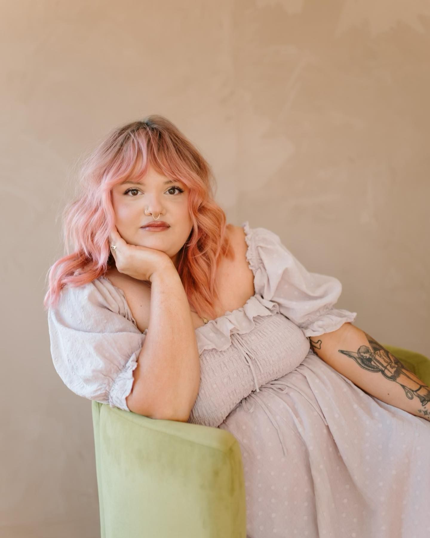 A woman with pink hair is sitting in a chair with her hand on her chin.