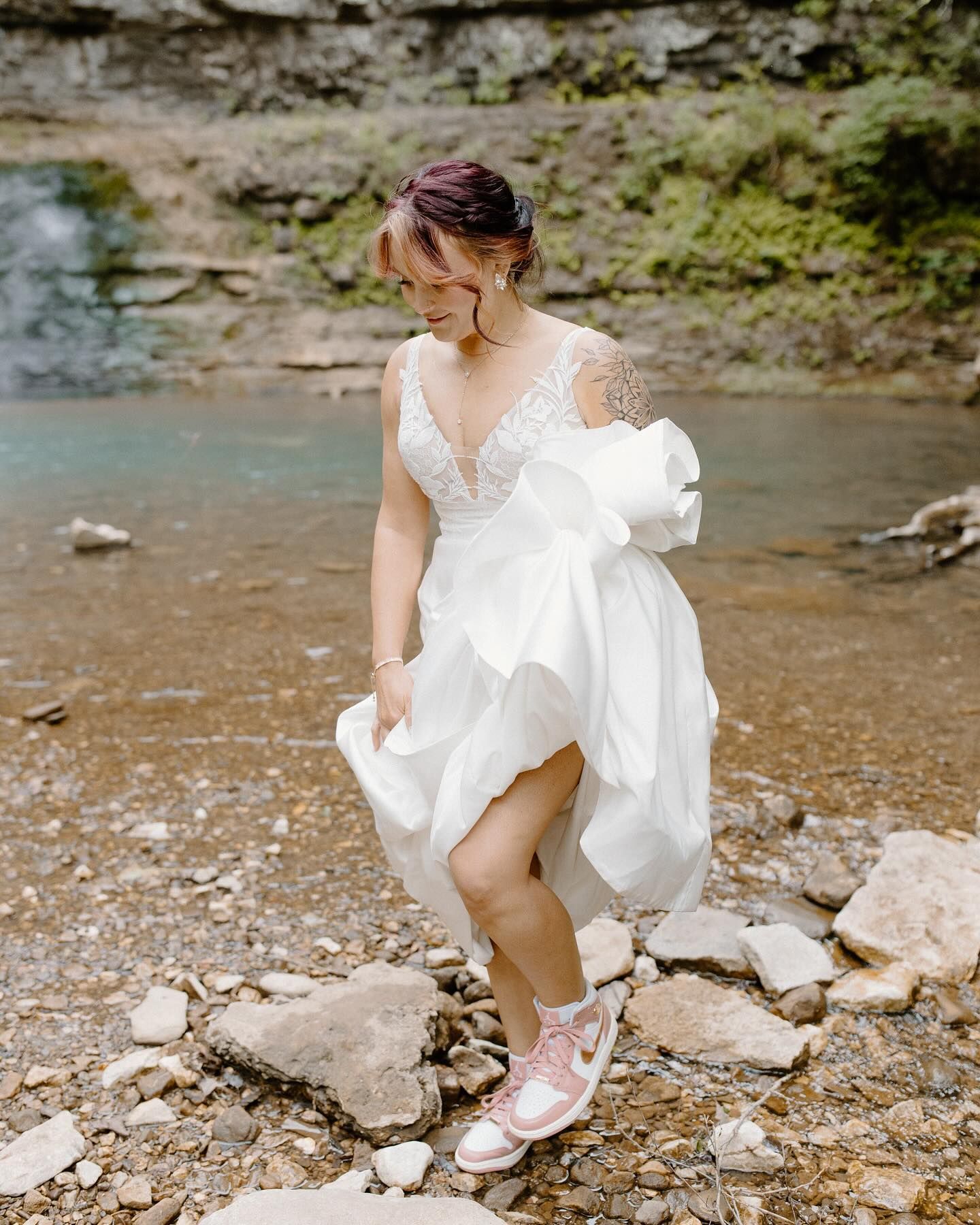 A bride in a wedding dress and pink sneakers is walking through a stream.