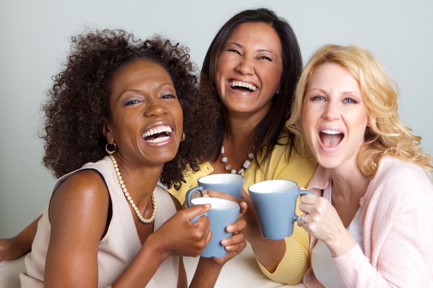 Three women laughing and holding mugs, indoors.
