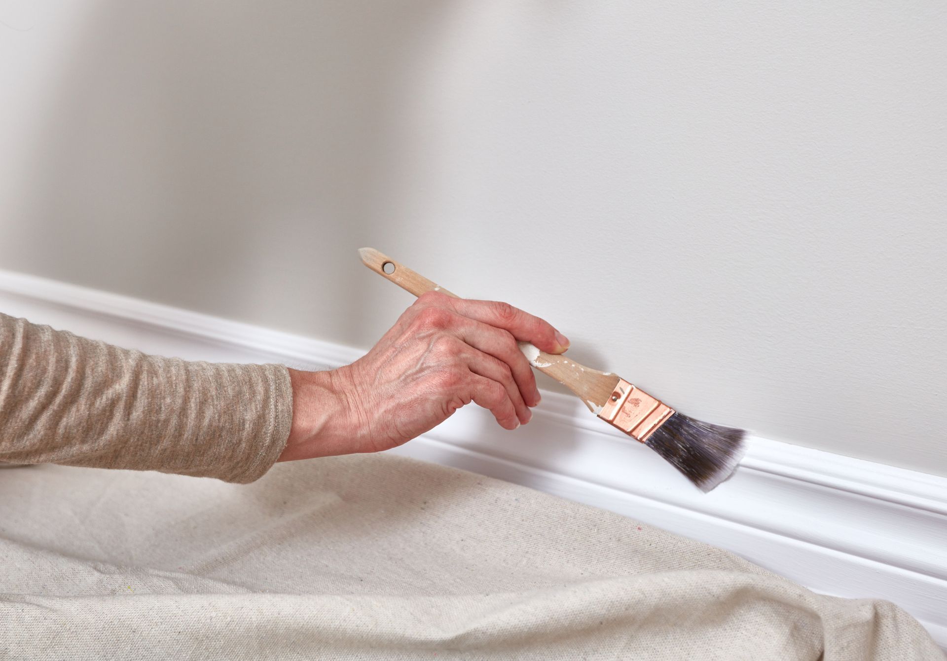 Hand painting white trim with a brush near a beige drop cloth.