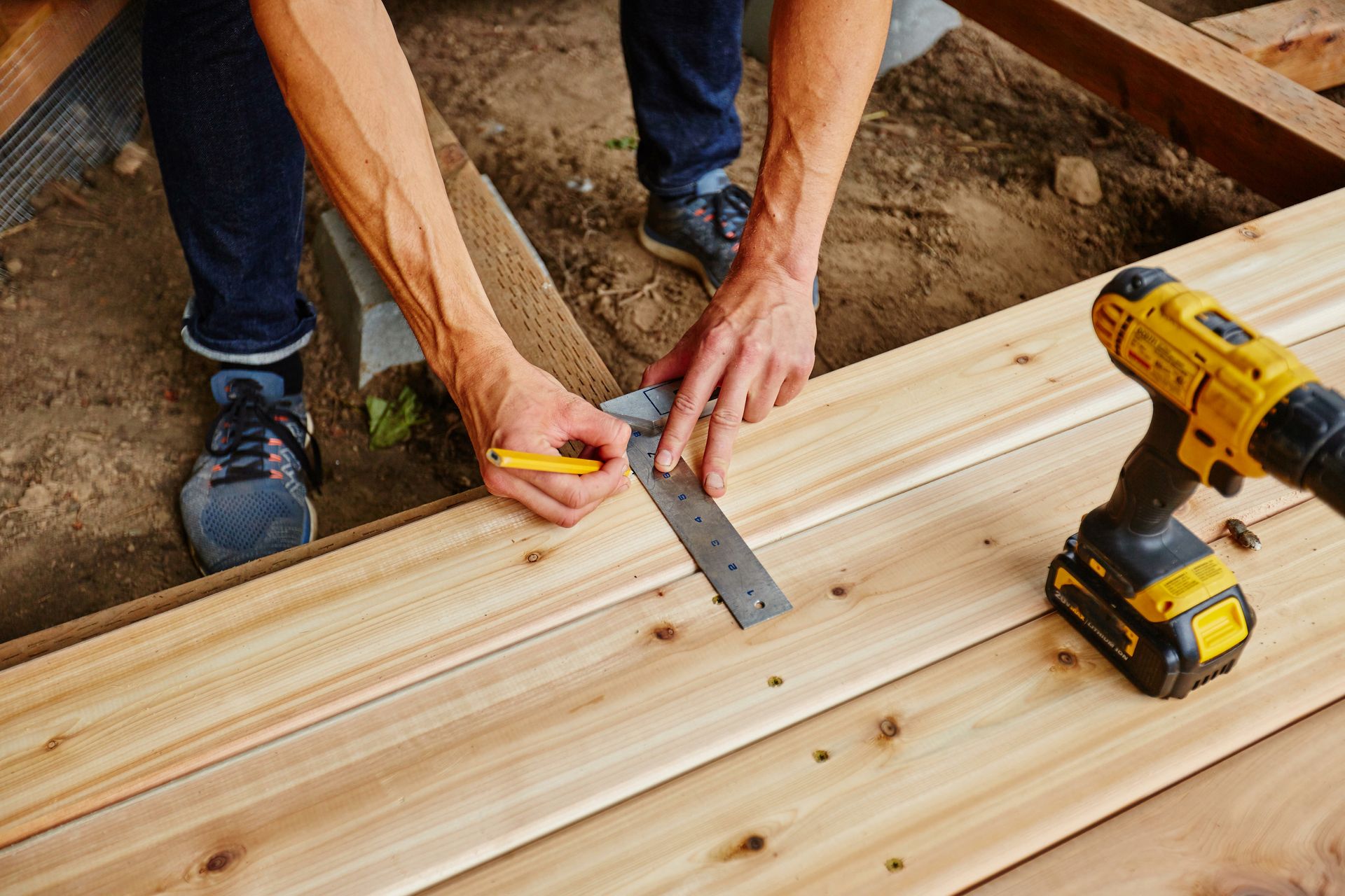 Person using a square to measure and mark wood planks, preparing to build a deck; a drill is nearby.