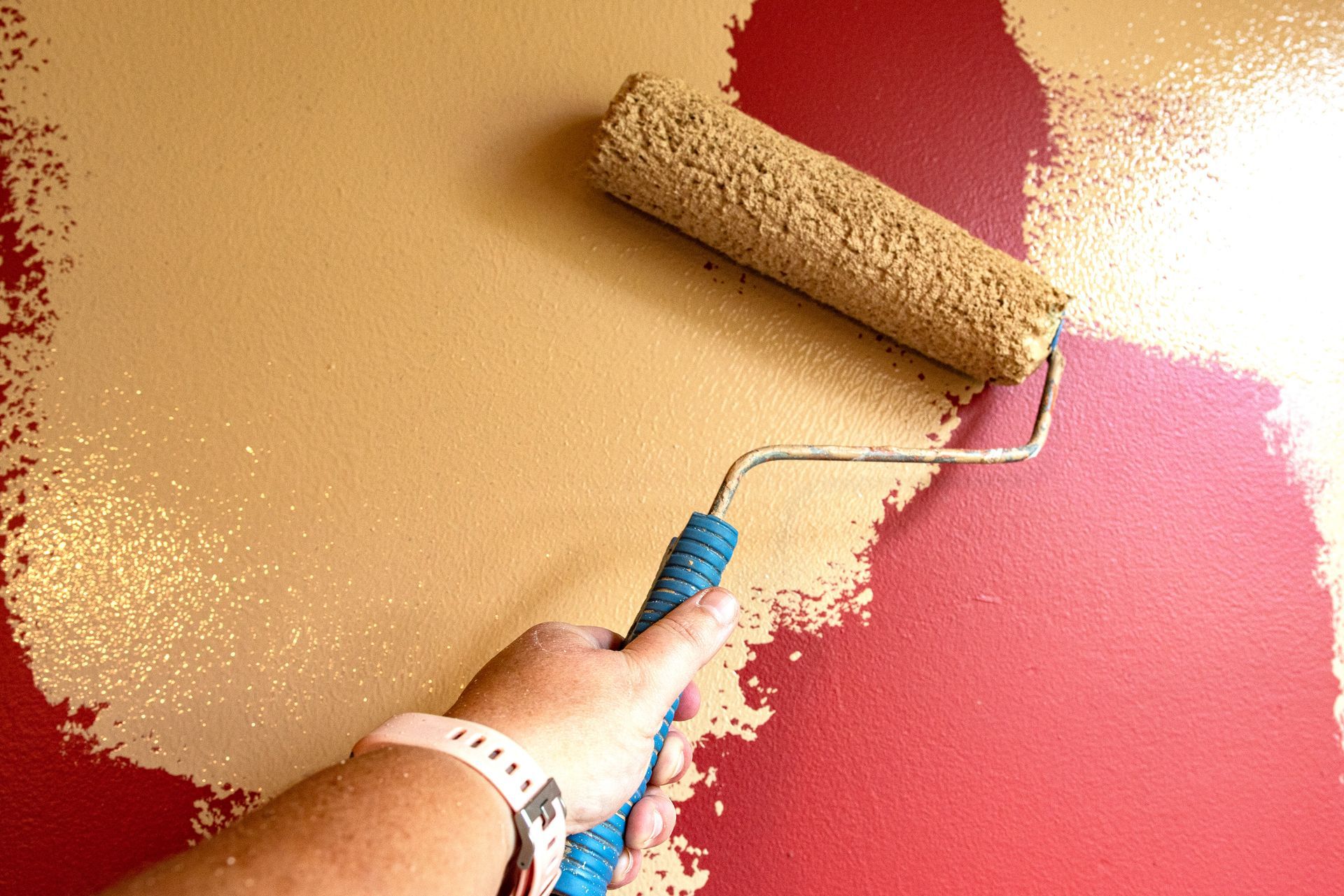Hand using a paint roller with tan paint, over a red painted wall.
