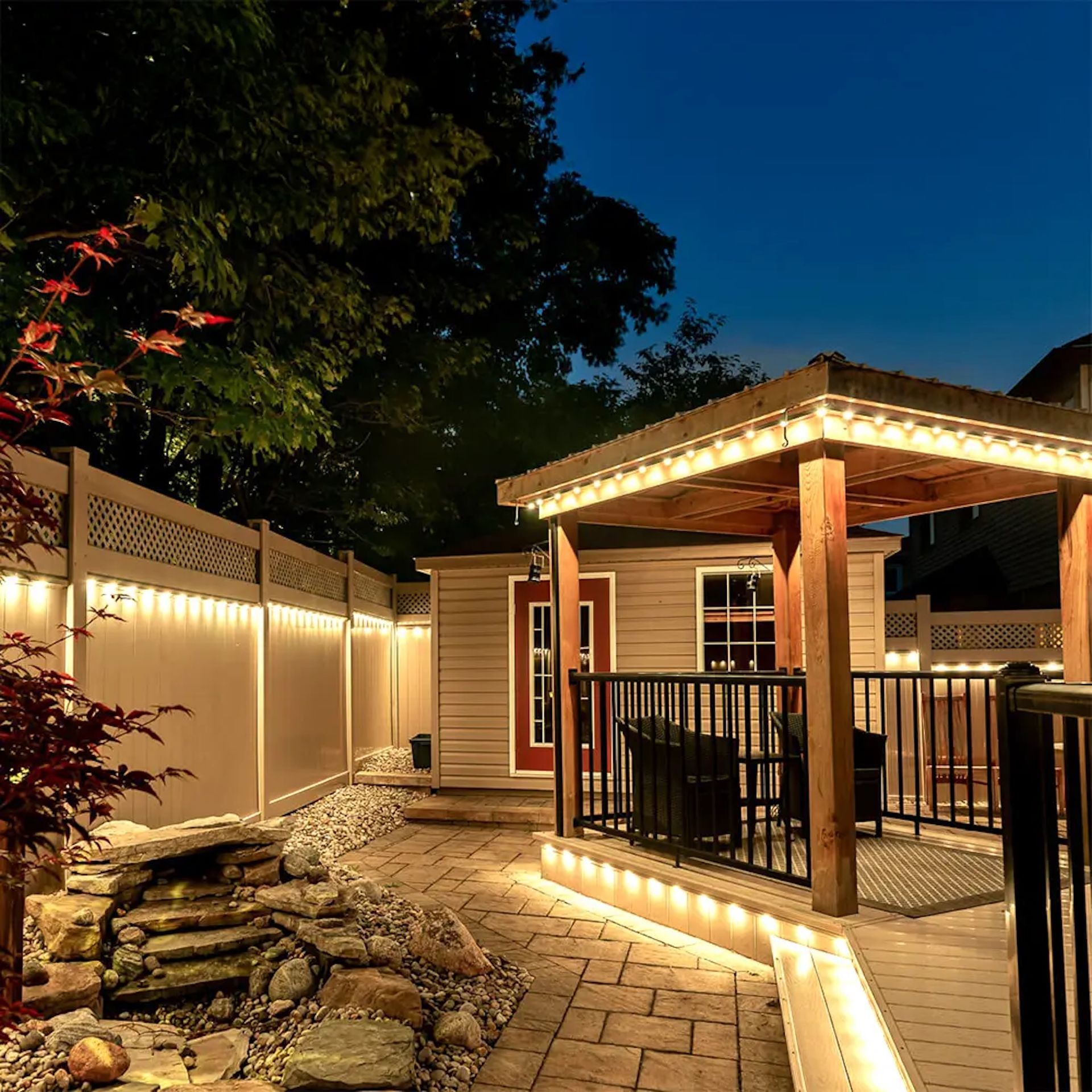 An illuminated backyard patio at night, featuring a gazebo, stone paving, a rock garden, and a small shed.
