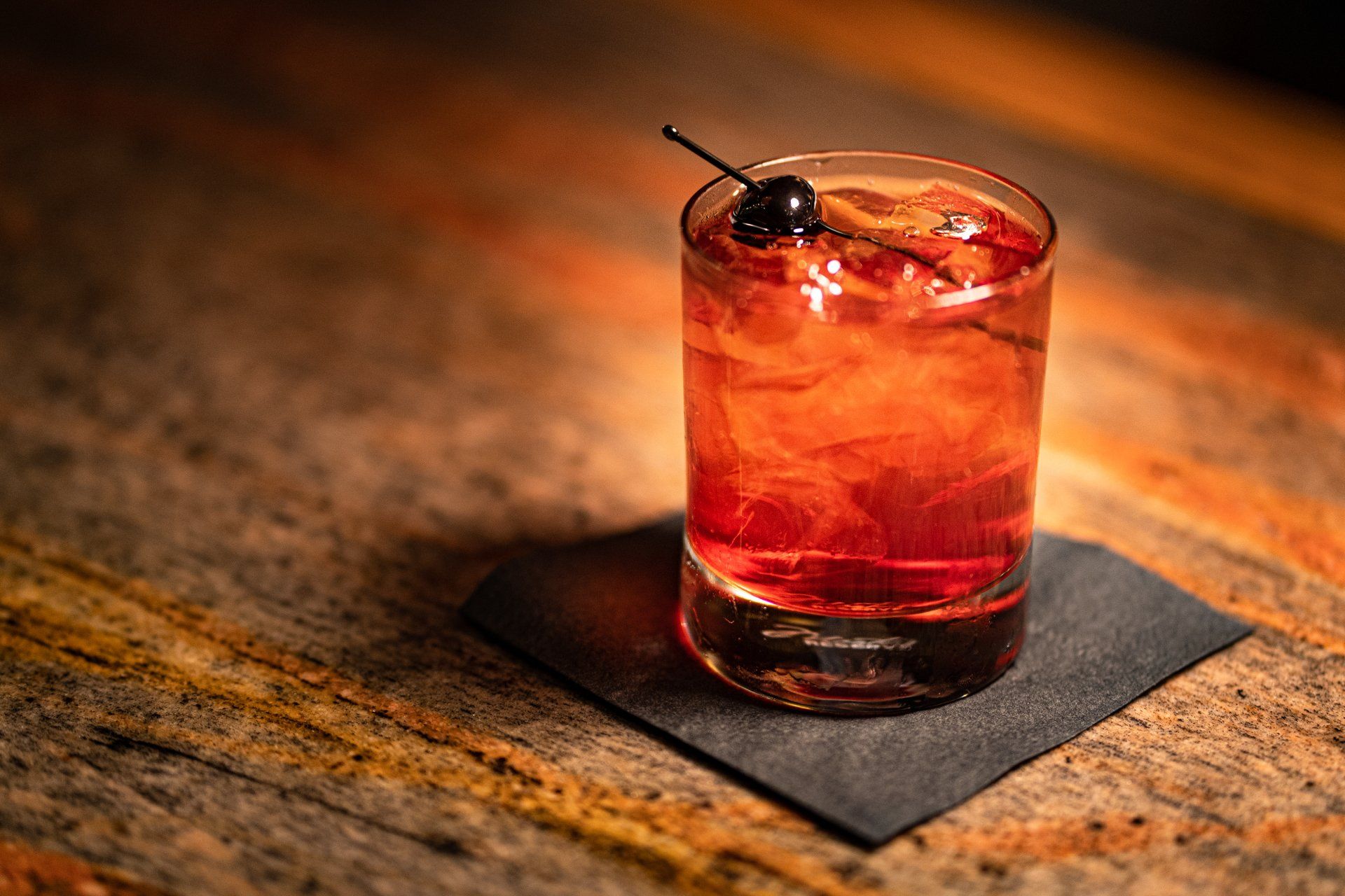 A glass of red liquid with ice and a cherry on a napkin on a table.