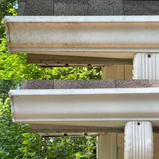 Before and after comparison of a white gutter being cleaned of dark oxidation stains against a background of green trees.