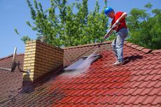 A person in a blue hard hat pressure washes a red tiled roof next to a brick chimney under a clear sky.